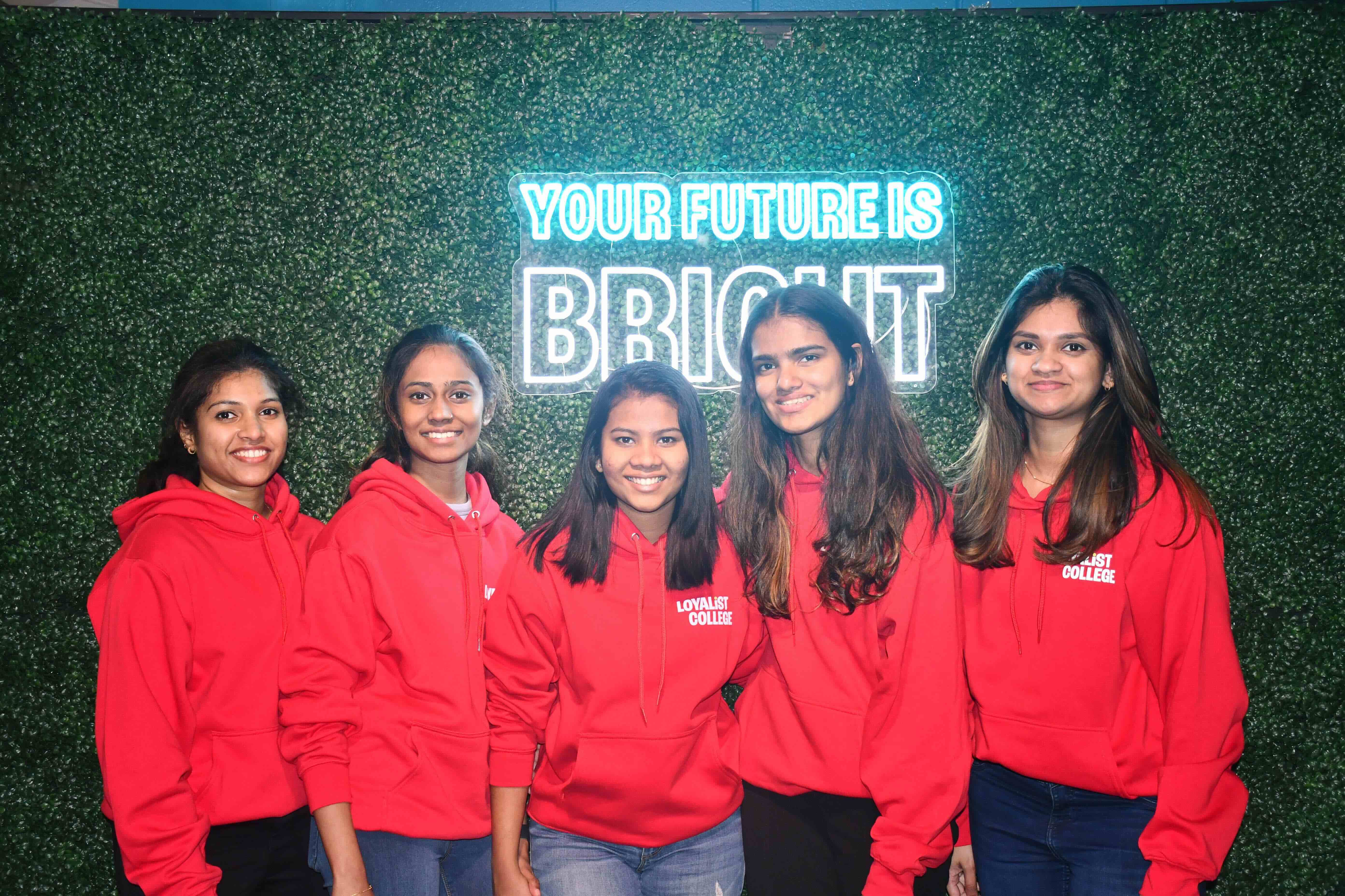 Alt text: Five young women wearing matching red Loyalist College hoodies stand together, smiling, in front of a green foliage wall with a neon sign that reads "Your Future is Bright." The group is posed confidently, conveying a sense of optimism and camaraderie.