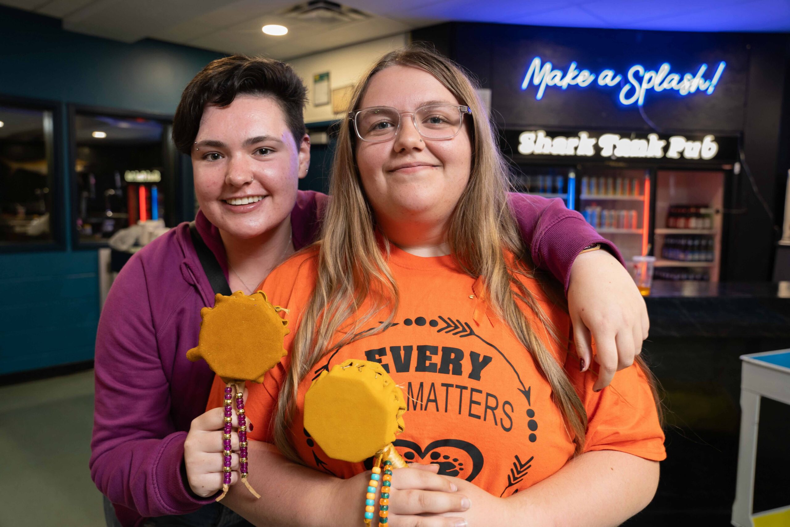 Two students are participating in a workshop during Truth and Reconciliation Week at Loyalist College. One student is wearing an orange "Every Child Matters" shirt, symbolizing their support for the cause. They both hold small, handmade drums decorated with colorful beads, reflecting their engagement in an Indigenous cultural activity. The workshop takes place in the Shark Tank Pub, creating a welcoming environment that emphasizes community, learning, and the importance of reconciliation.