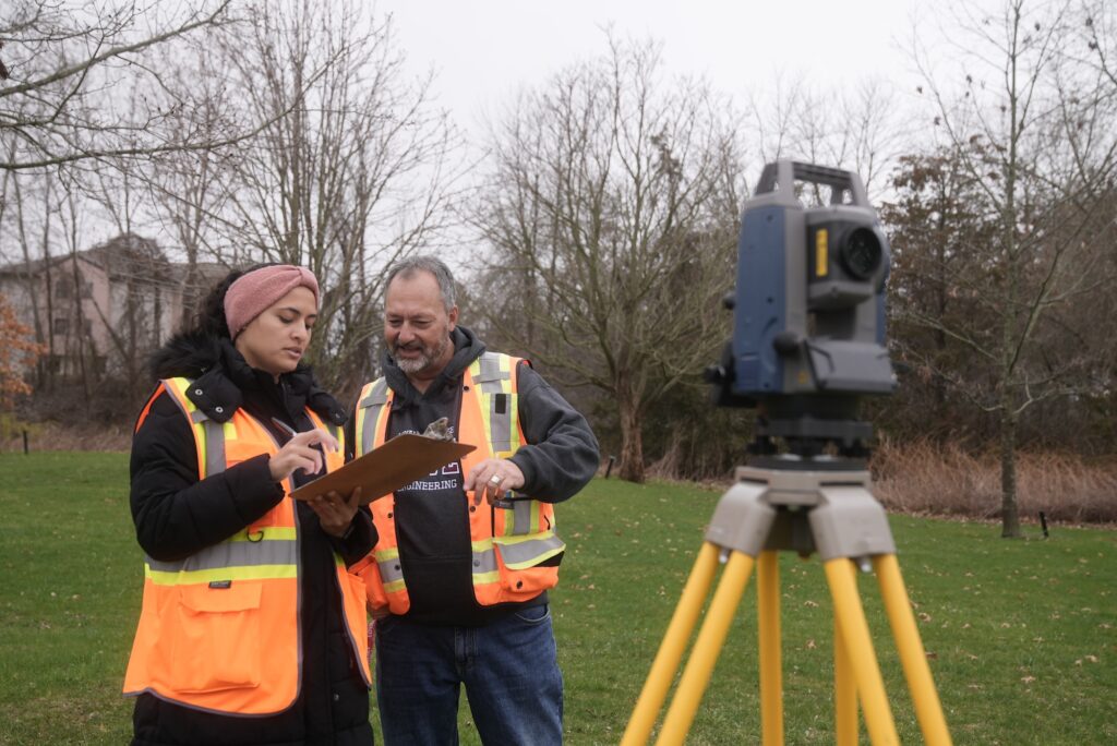 A student and faculty member at Loyalist College engaged in fieldwork, both wearing orange and yellow construction neon vests. They are examining a clipboard, suggesting a focus on surveying and engineering technician activities.