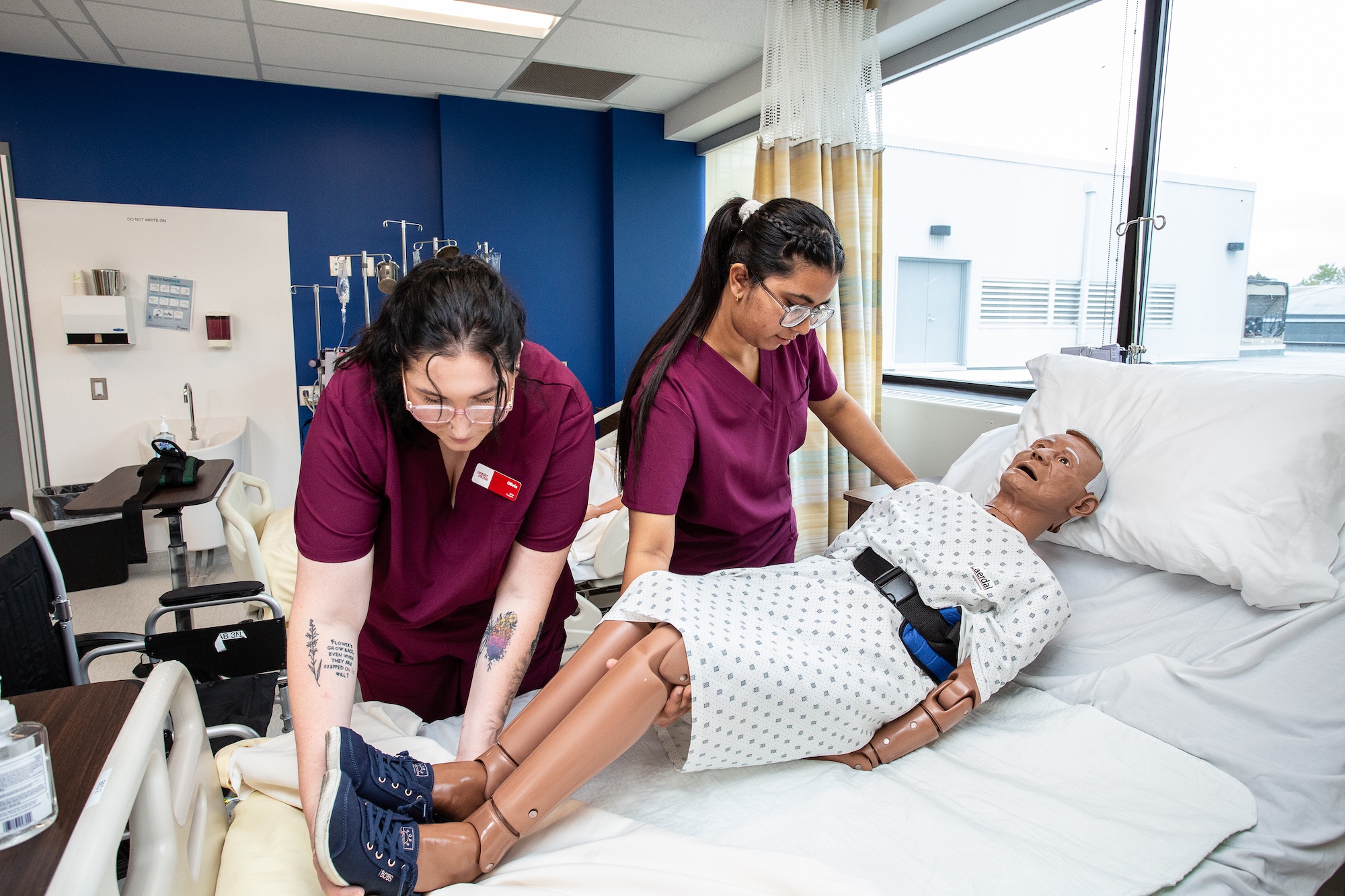 Two students wearing purple scrubs are working with a mock patient laid down on a bed.