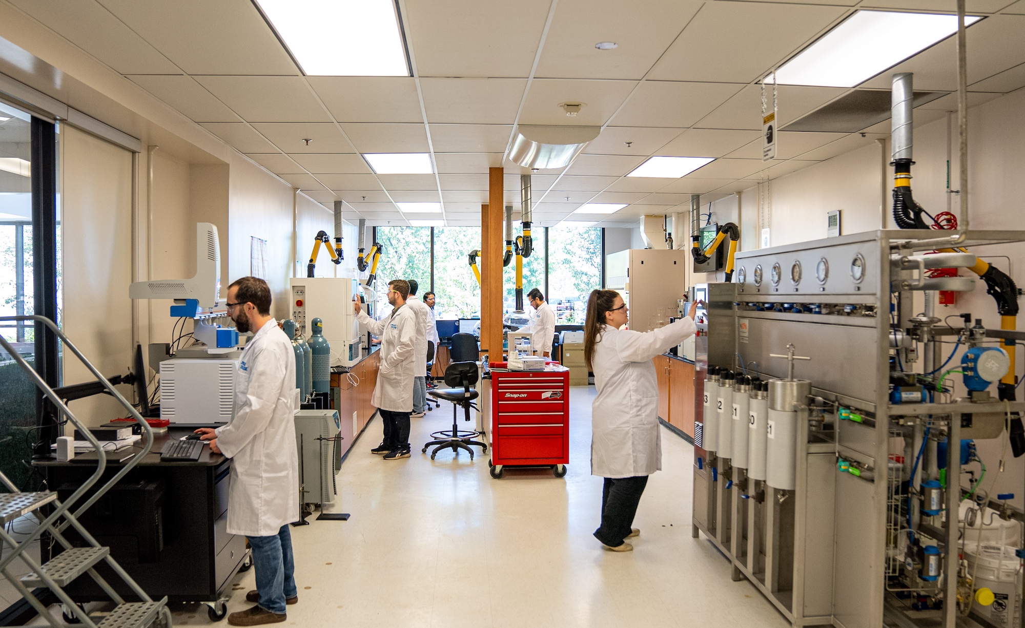 A group of people wearing white lab coats and a pair of goggles are scattered in different areas of a room working with devices and tools.