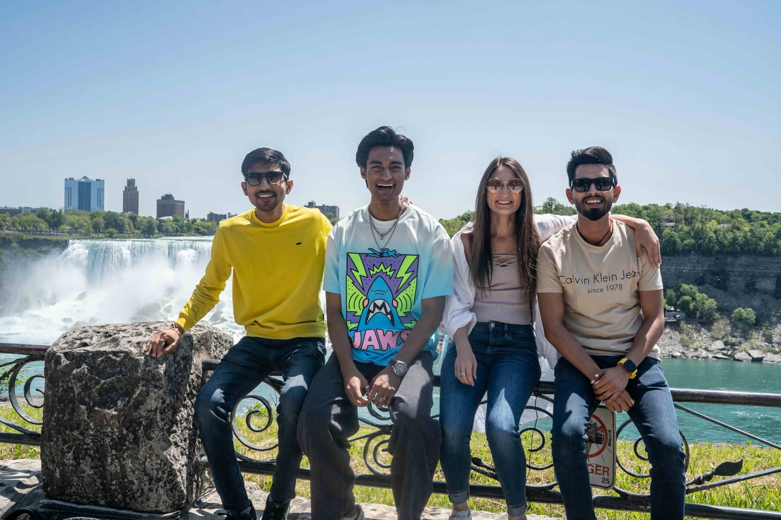 A group of four friends sitting on a railing with Niagara Falls in the background, enjoying a sunny day. They are smiling and relaxed, capturing the moment with the iconic waterfall and cityscape behind them.