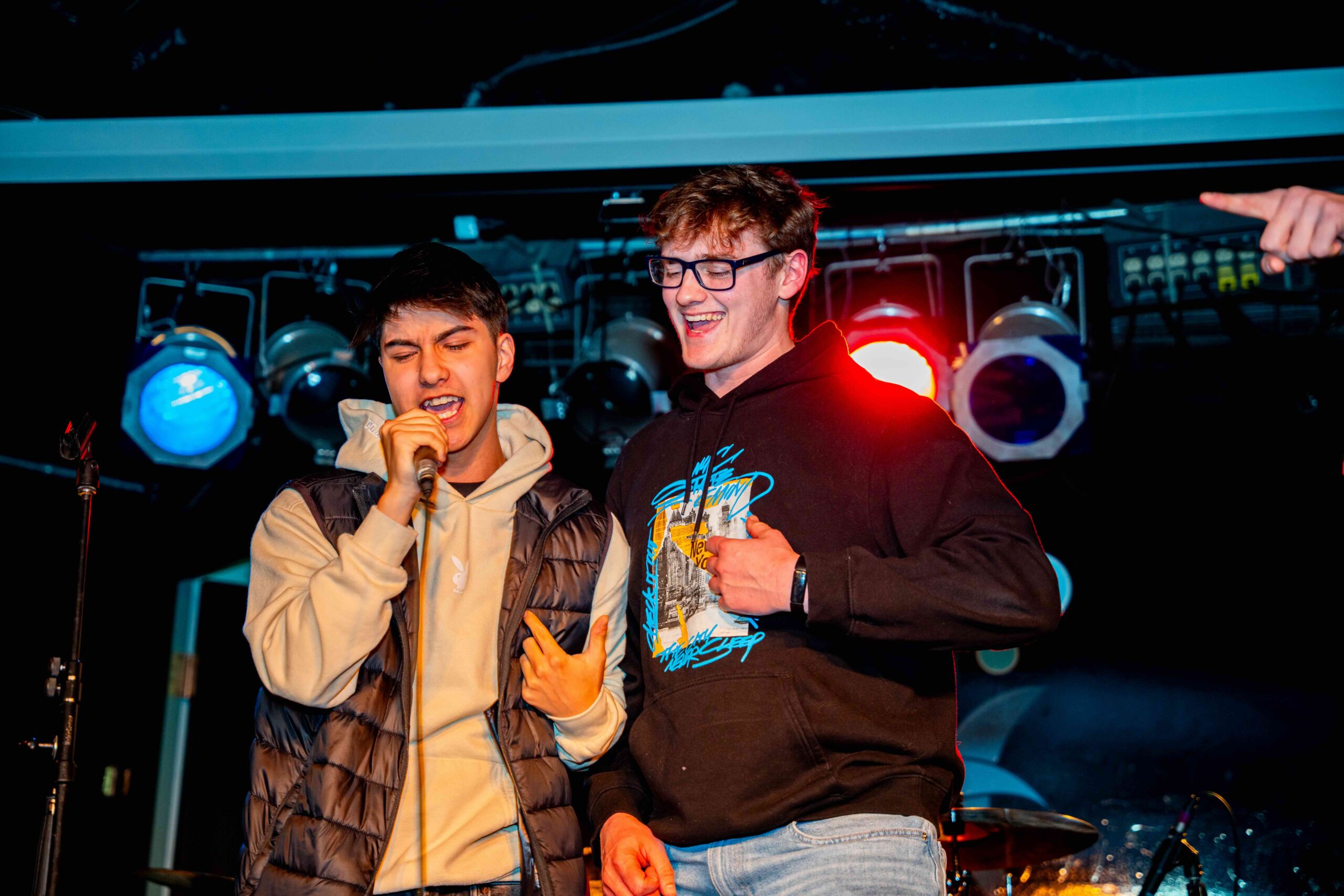 Two students are on stage during a karaoke event, one passionately singing into a microphone while the other laughs and supports him. The scene is lit with colorful stage lights, creating a lively and energetic atmosphere.