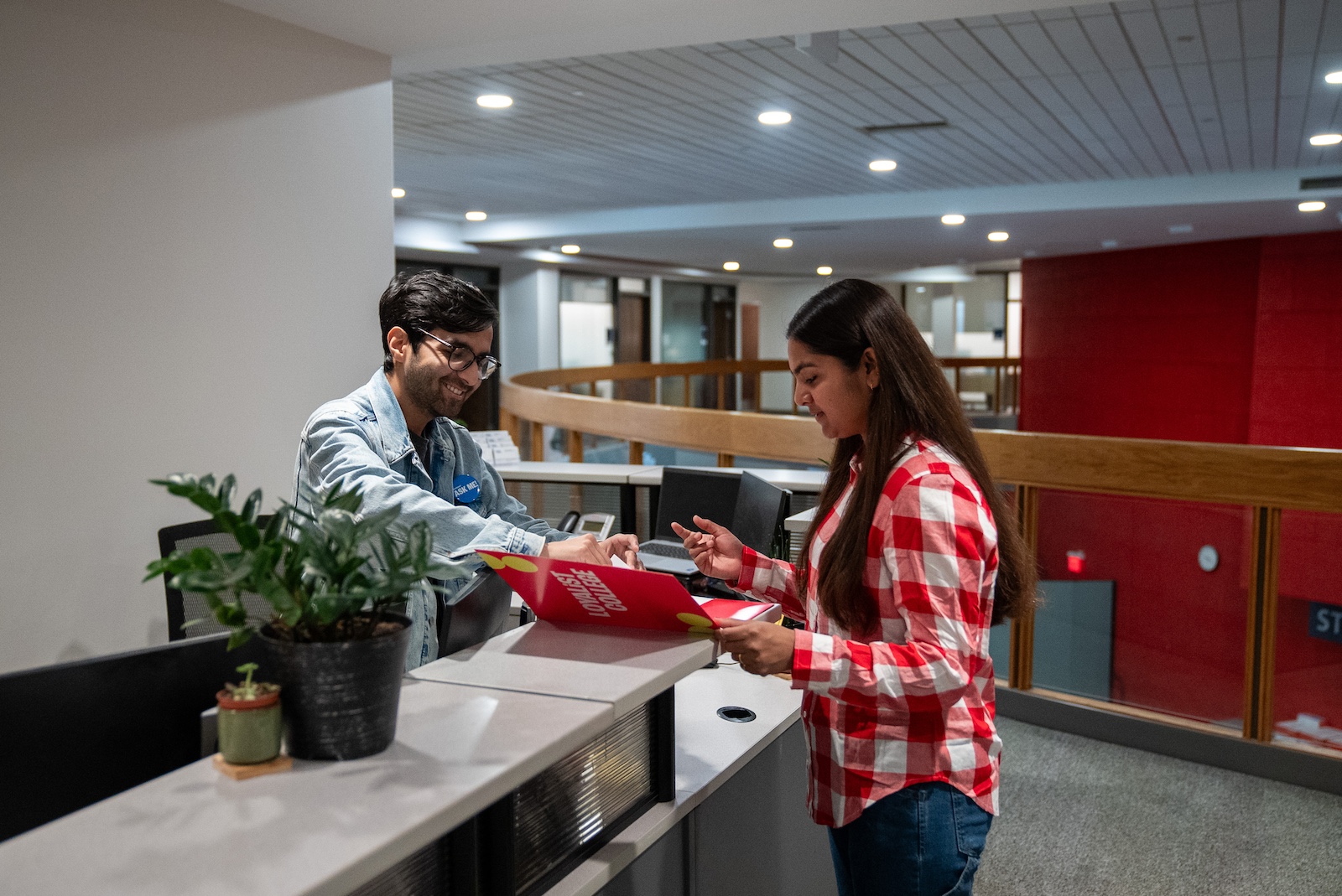 Two students stand on either side of a reception desk. One student is showing another the contents of a red folder that says "Loyalist College" on the front.