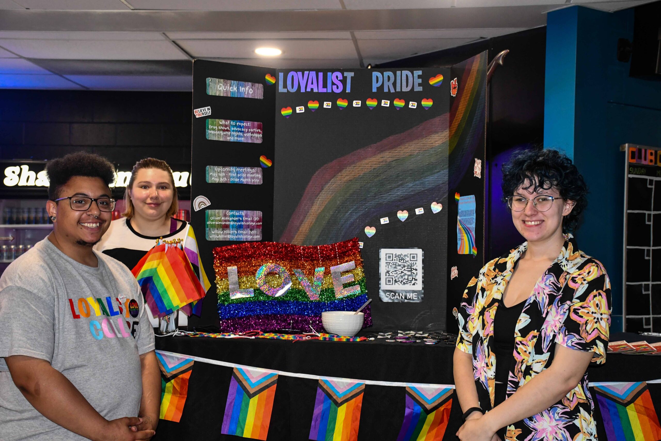 Three students standing proudly beside a vibrant and colorful 'Loyalist Pride' display booth, decorated with rainbow flags and a sparkling 'LOVE' sign. The booth is set up to promote inclusivity and LGBTQ+ awareness, with informational posters and a QR code for more details.