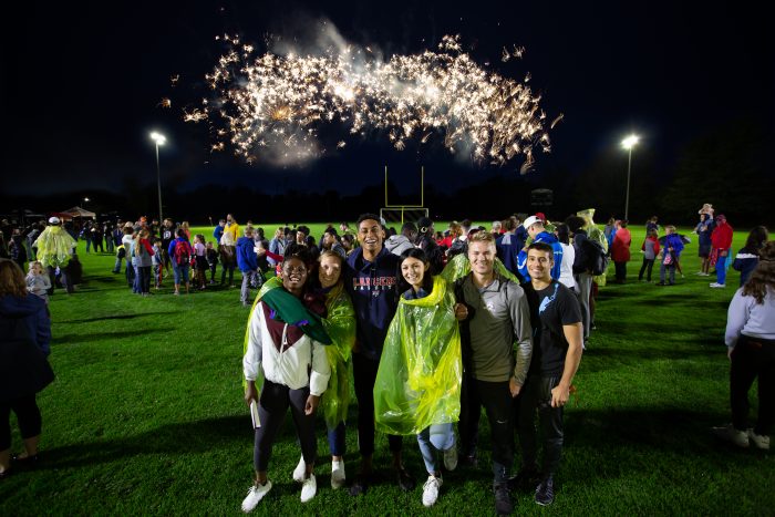 Students are gathered around a football field at night. A small group of them smiles into the camera with fireworks in the background.