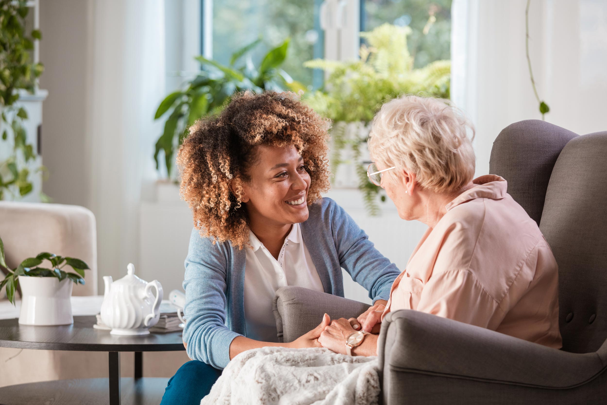 A young person crouches beside a large armchair, holding the hands of an older person with grey hair and glasses.