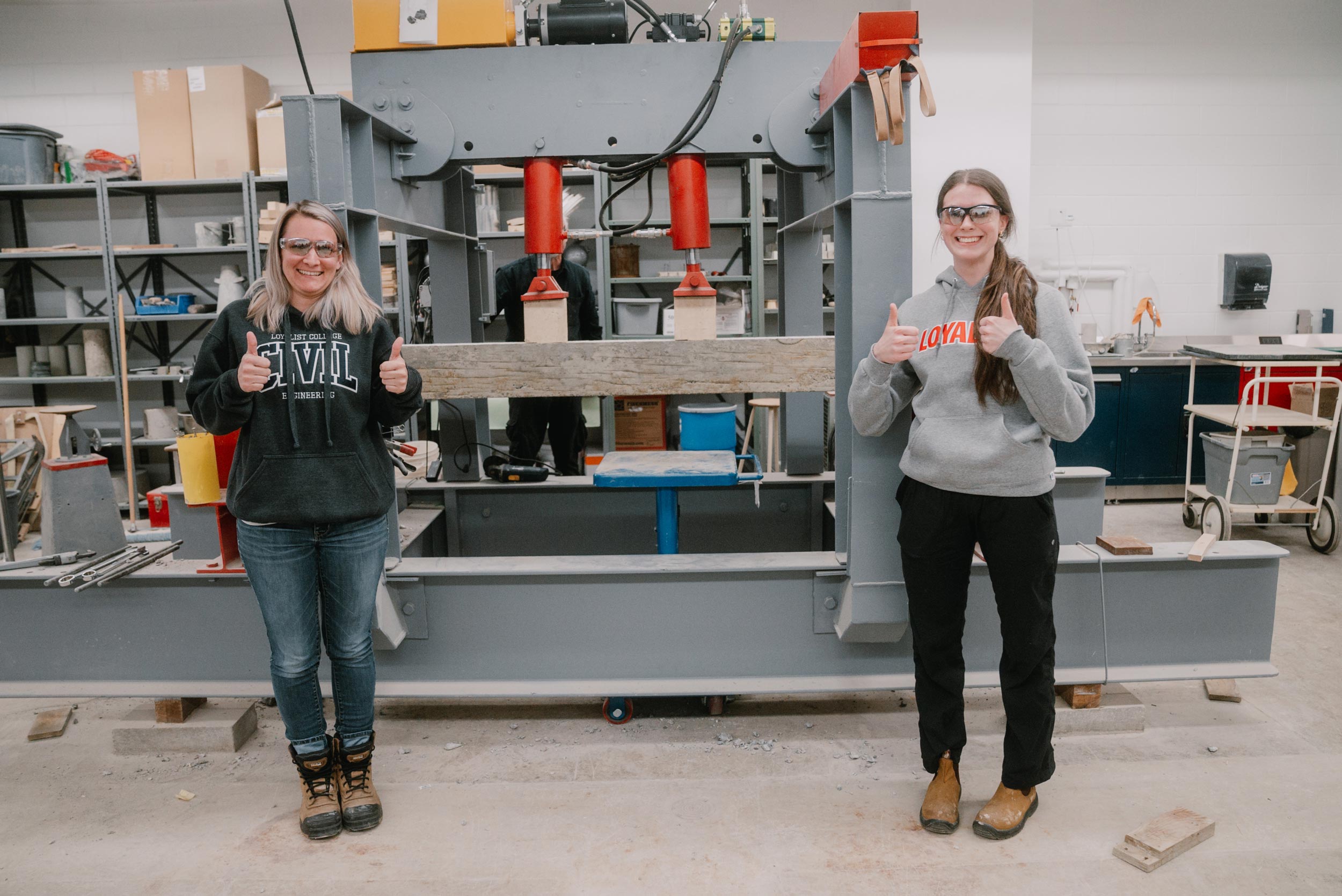 Two women in a workshop give thumbs up while standing in front of a large hydraulic press machine. The woman on the left wears a "Loyalist College Civil Engineer" hoodie, and the woman on the right wears a "Loyalist" hoodie. Both are smiling, wearing safety glasses, and standing on either side of a piece of wood being tested by the machine.