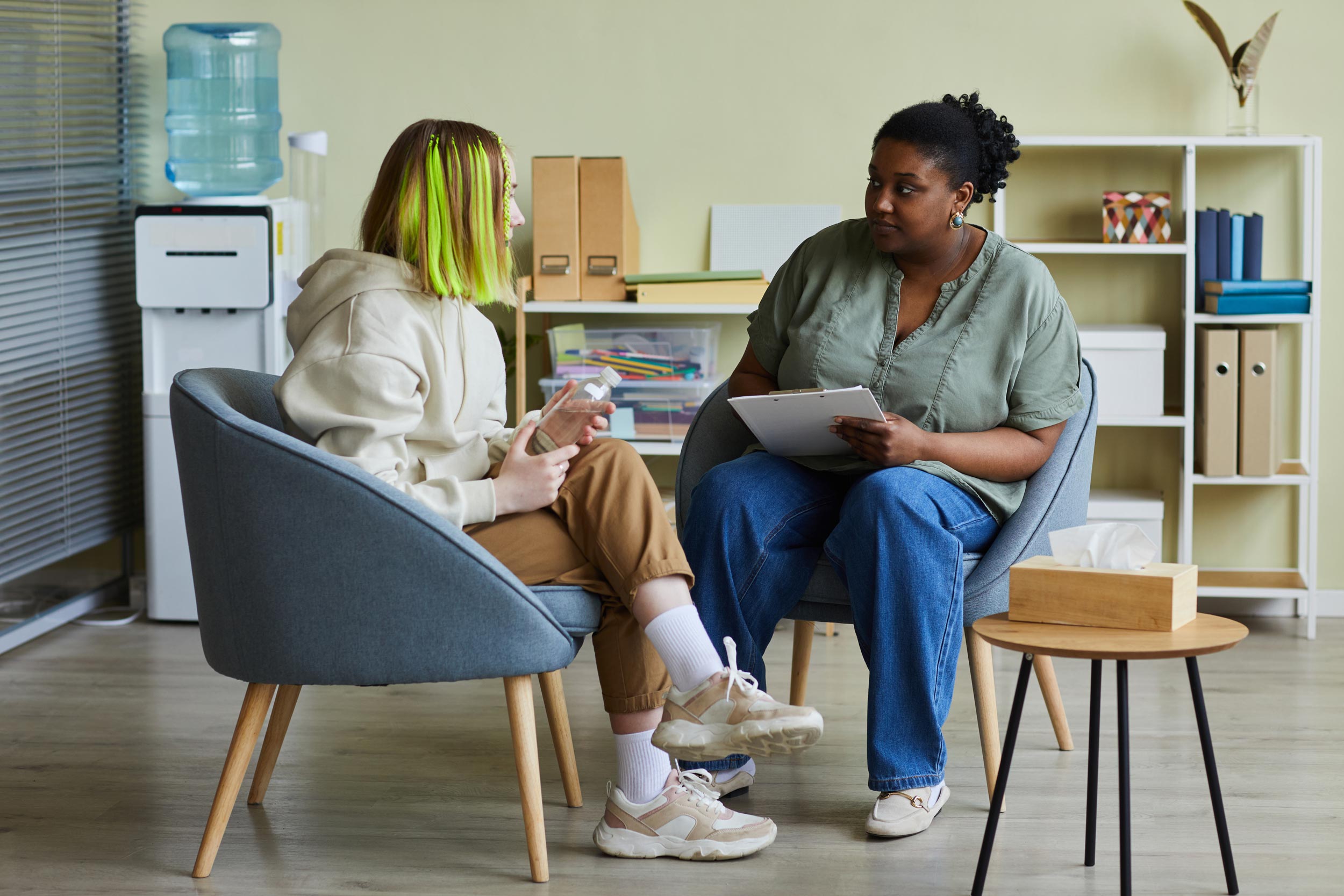 A young person with vibrant green hair sits in a chair holding a water bottle, engaging in conversation with a counselor or social worker, who is holding a clipboard. They are in a cozy, well-lit office space with a water dispenser, bookshelves, and a small table with a tissue box.