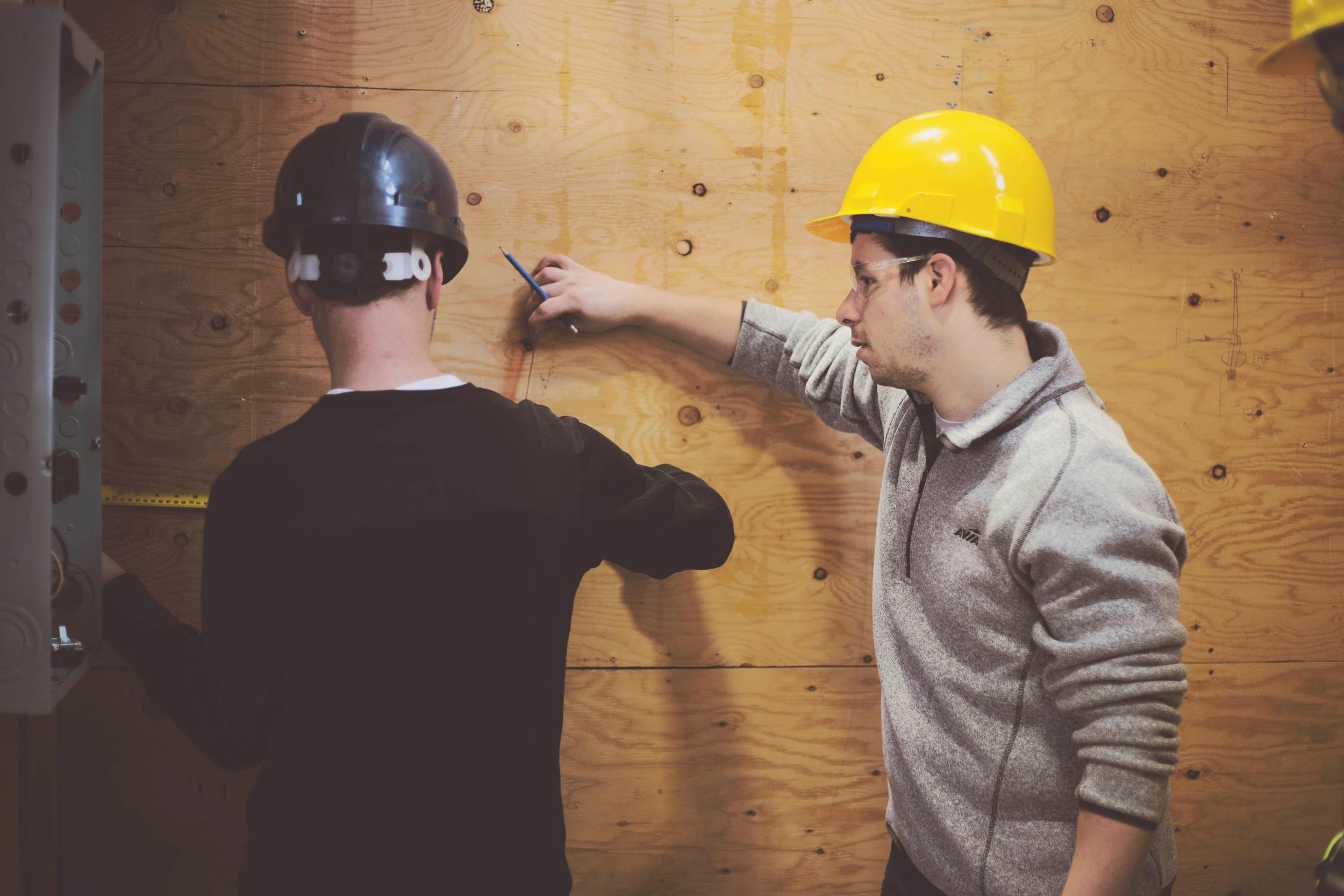 Two students are standing side-by-side each other. The students are wearing hard hats. One is using a measuring tape against a wooden board and the other person is leaning their wrist on the wall holding a pencil.