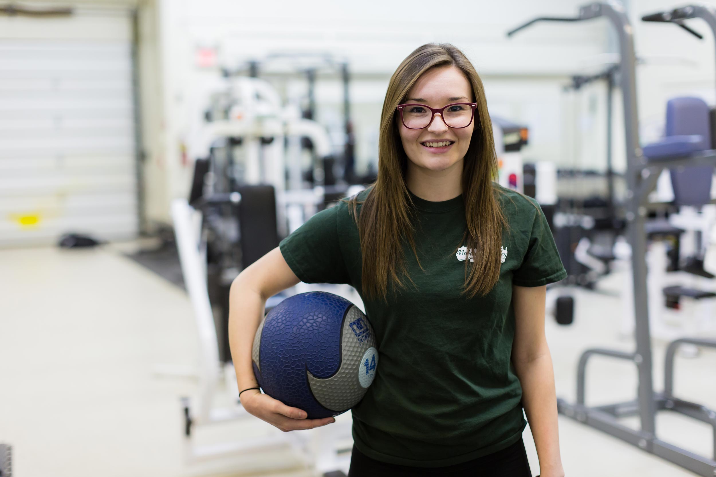 A student in the fitness and health promotion program is smiling towards the camera holding a medicine ball in one hand.