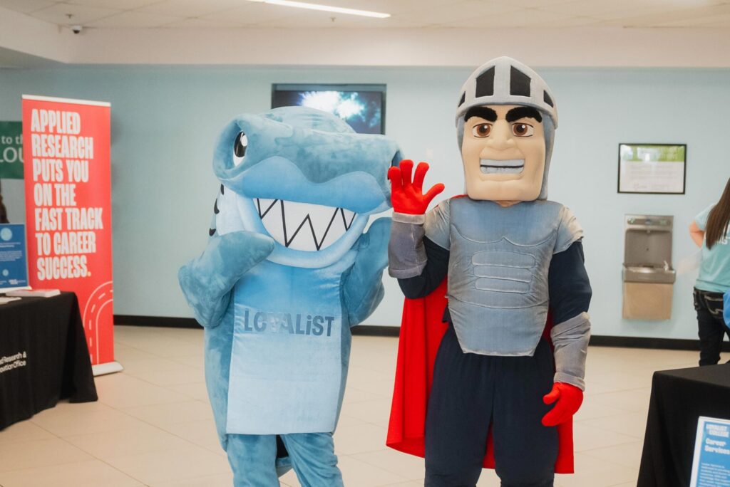 Loyalist mascots Lenny who looks like a knight and Sharkie who is a blue shark stand together at an open house. A sign in the background says "Applied research puts you on the fast track to career success."