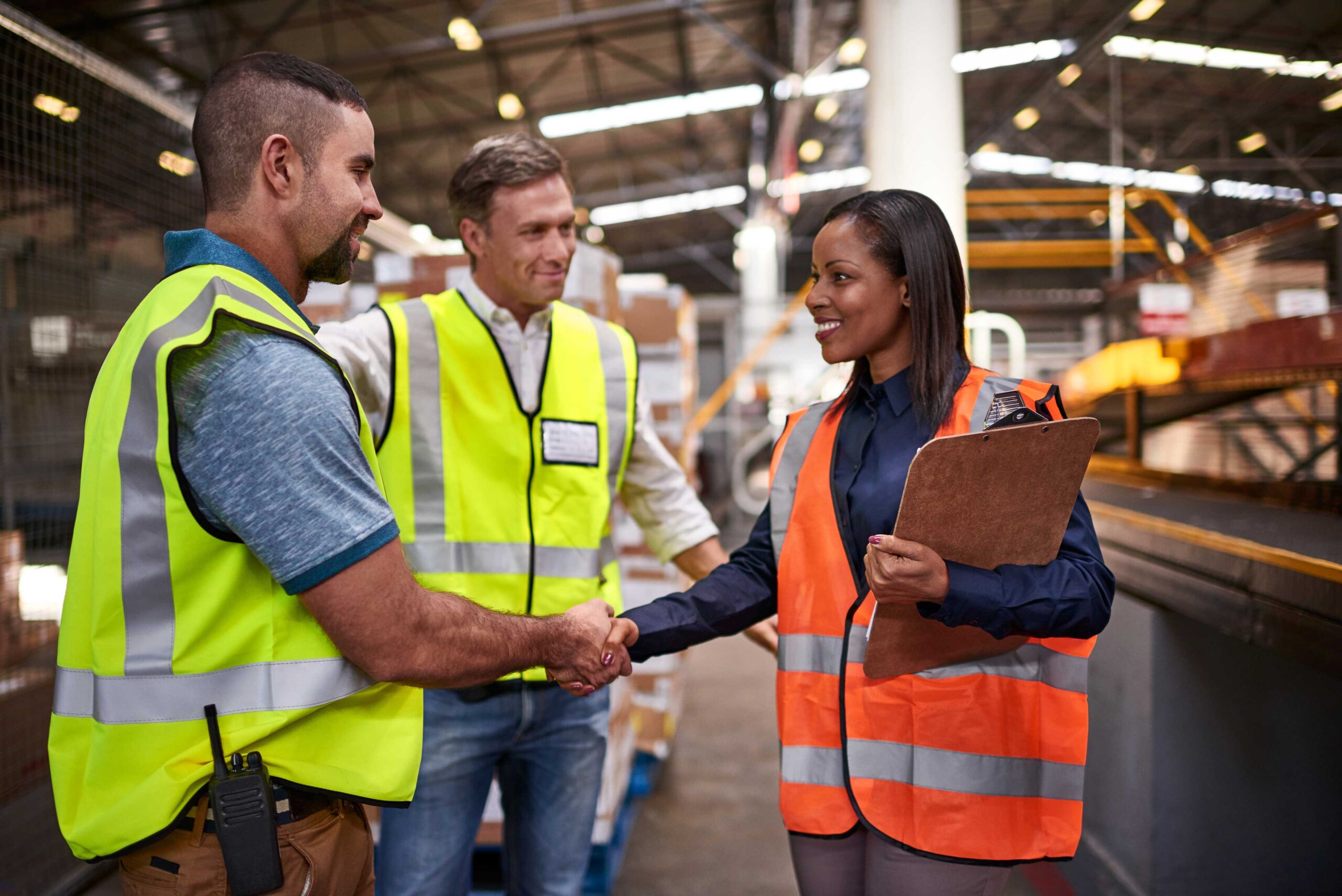 Two people in high visibility vests shake hands in an industrial warehouse. One holds a clipboard. Another person in a high visibility vest stands beside them.