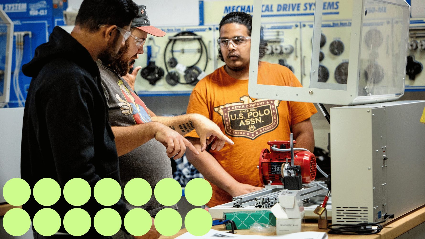 Two students listen to the professor, who points to a piece of mechanical equipment on a workbench. All three are wearing safety glasses. Green graphic circles are in the lower lefthand corner of the image.