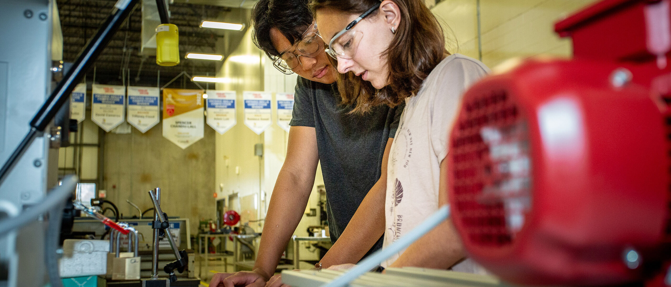 Two students work side-by-side at a workbench full of mechanical equipment. Both are wearing safety glasses.