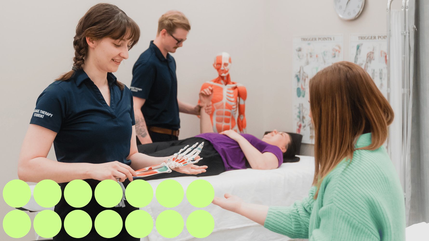 A massage therapy student shows a skeletal model of a hand to another person holding out a hand. In the background, another massage therapy student holds the wrist of a person lying on a massage table. Green graphic circles are in the lower lefthand corner of the image.