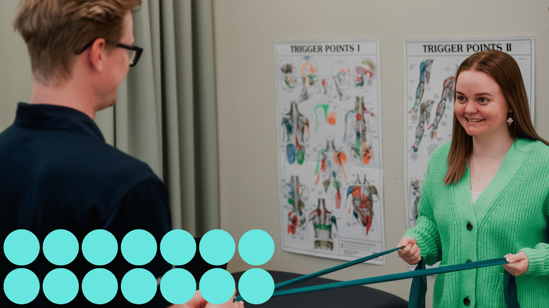 A massage therapy student holds an exercise band while another person pulls on the other end, smiling and standing in front of two posters containing trigger point diagrams. Blue graphic circles are in the lower lefthand corner of the image.