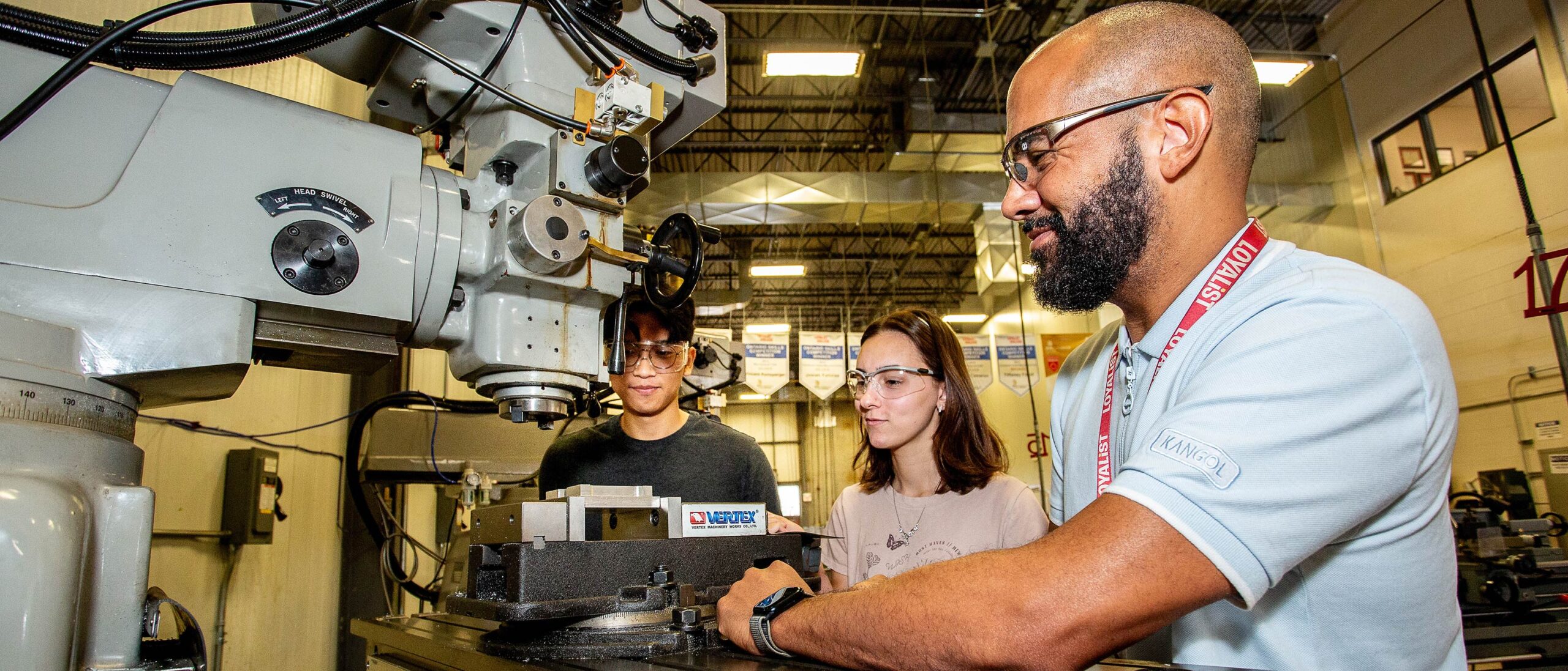 A professor and two students in the Mechanical Techniques program are smiling as they examine a tool together in a classroom setting. They are engaged in a discussion, fostering a collaborative learning environment.