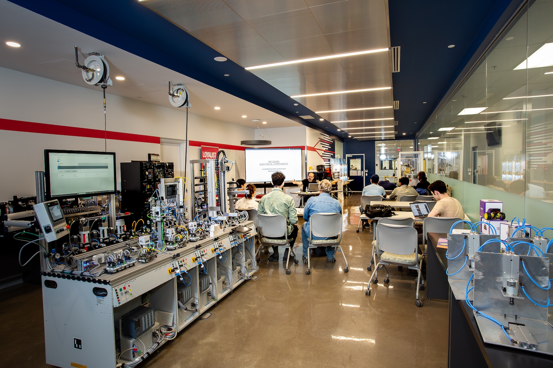 Students sit at desks listening to a professor teaching in front of a presentation. Behind the students is mechatronics equipment.