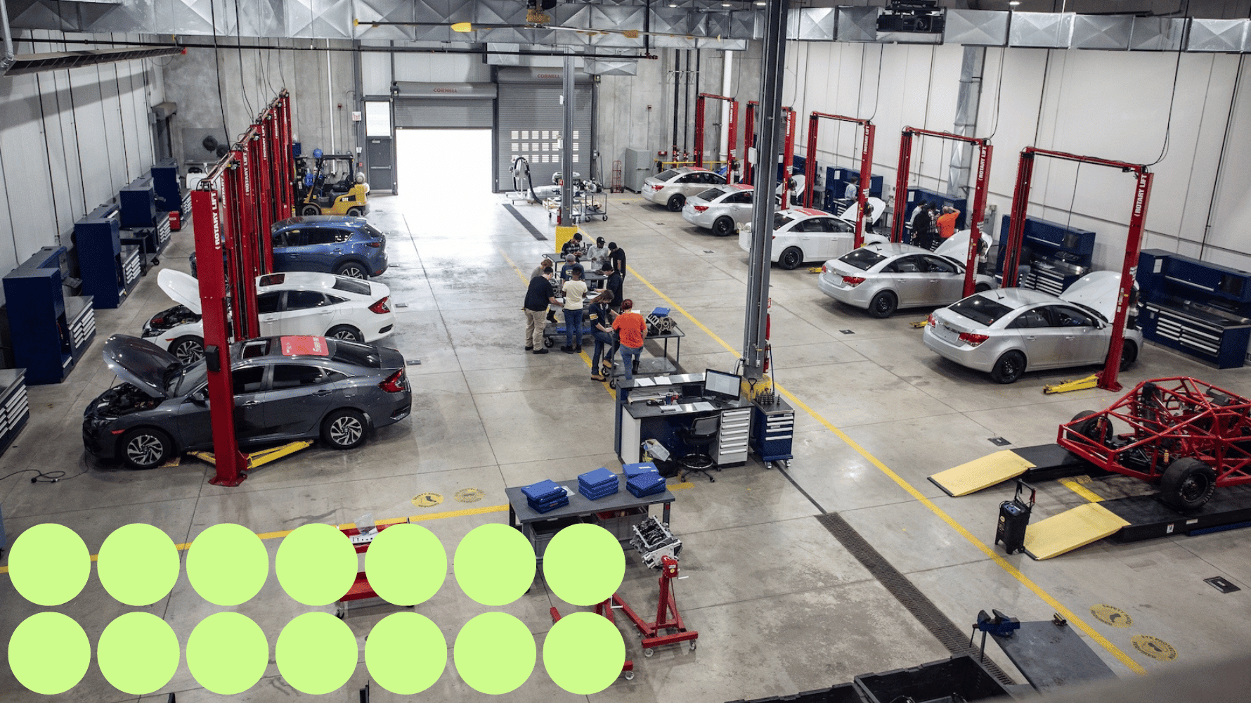 A group of motive power fundamentals students are standing inside their classroom surrounded by car lifts, cars, and automotive tools. Fourteen green graphic circles are in the lower lefthand corner of the image.