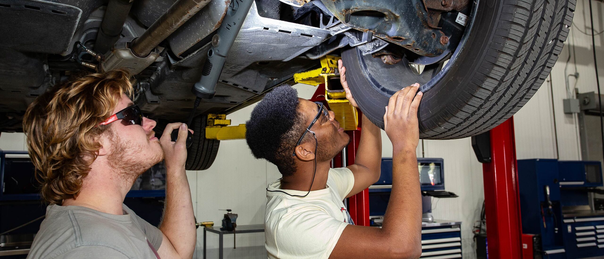 A close-up shot of two students working underneath a car while it's on an industrial car lift. The two student are wearing goggles and looking upwards to the car.