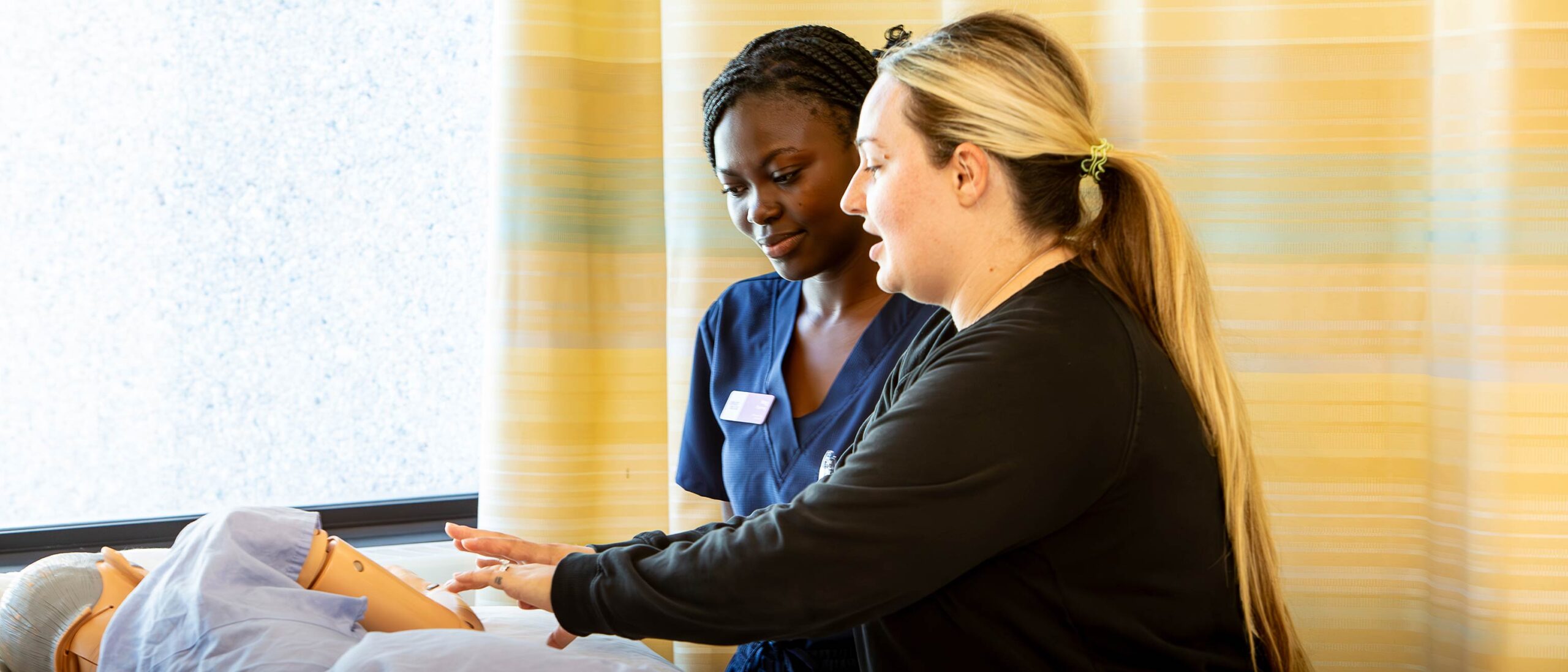 A professor is working with a student on instructing what to do with the mock patient inside a hospital room.