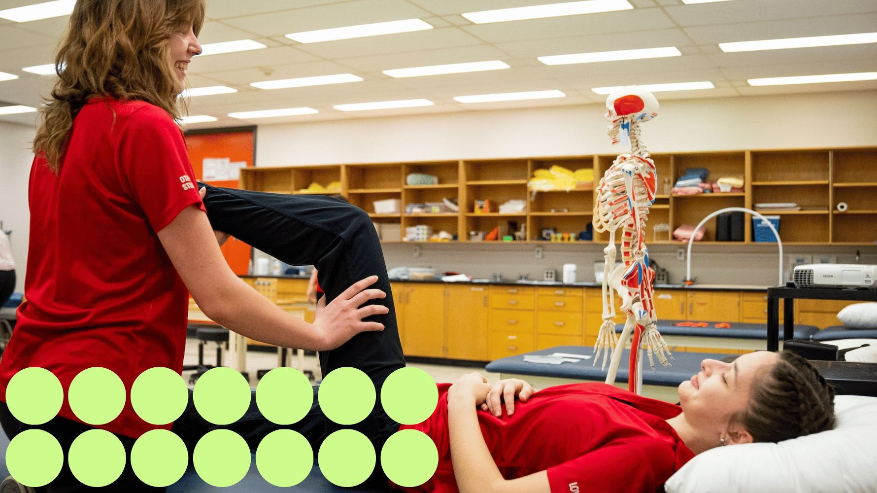 Two students are in the occupational therapist assistant and physiotherapist program. One student is helping stretch the other student's leg while they are laying down. They are both wearing bright red shirts. Fourteen green graphic circles are in the lower lefthand corner of the image.