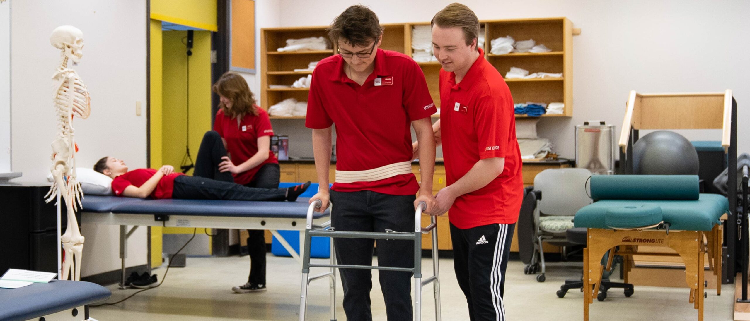 There are two scenarios happening in the image. In the forefront of the image, there are two students. One student is helping the other student walk. In the back left of the image, there is one student help stretch another student's leg while they are laid down. Everybody is wearing a bright red shirt in a mock clinic setting.