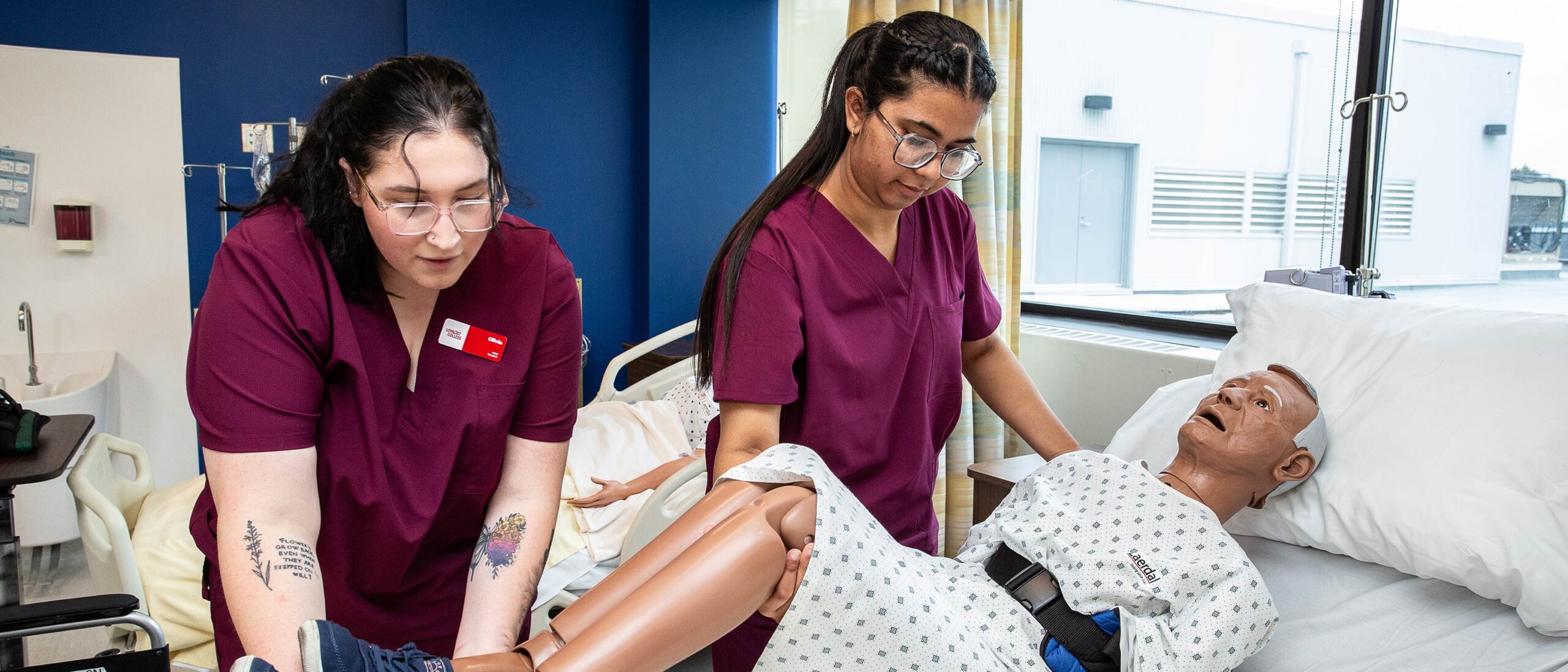 Two students in the personal support worker program is working with a mock patient laid down in a classroom clinic setting. The students are both wearing burgundy coloured scrubs.