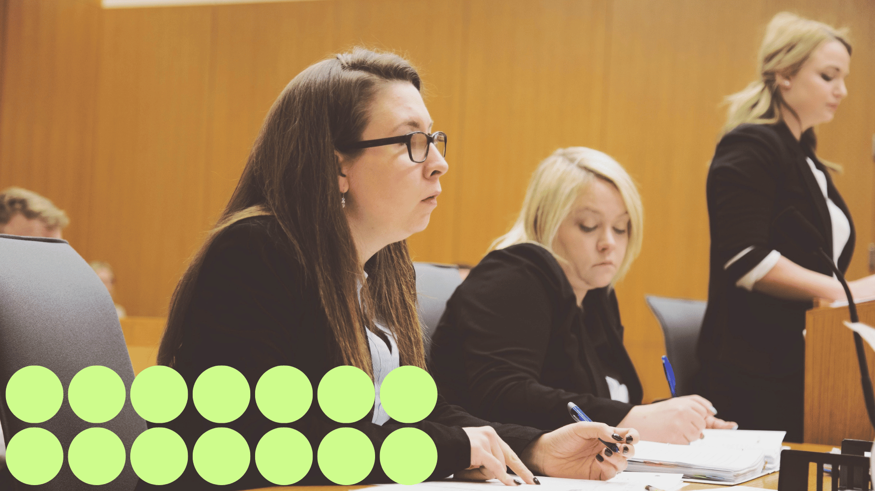 This image is depicting a paralegal scenario inside a courtroom. Two people are sitting down. One is looking towards the right of the image and one is writing down on a piece of paper. A third person is standing up looking down in front of them. There are also people sitting behind them observing. Fourteen green graphic circles are in the lower lefthand corner of the image.