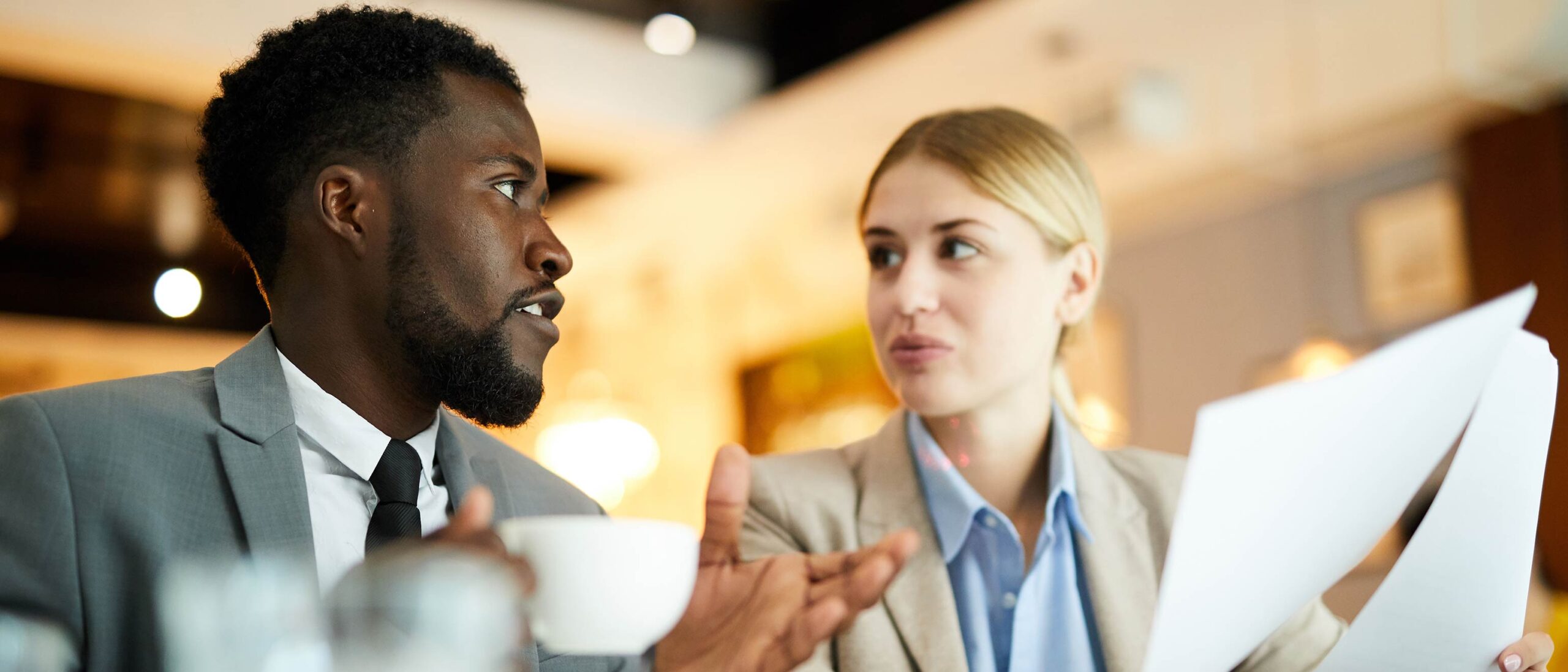 Two paralegal students are engaged in conversation, both wearing professional attire. One student is holding a piece of paper, while the other holds a white glass mug.