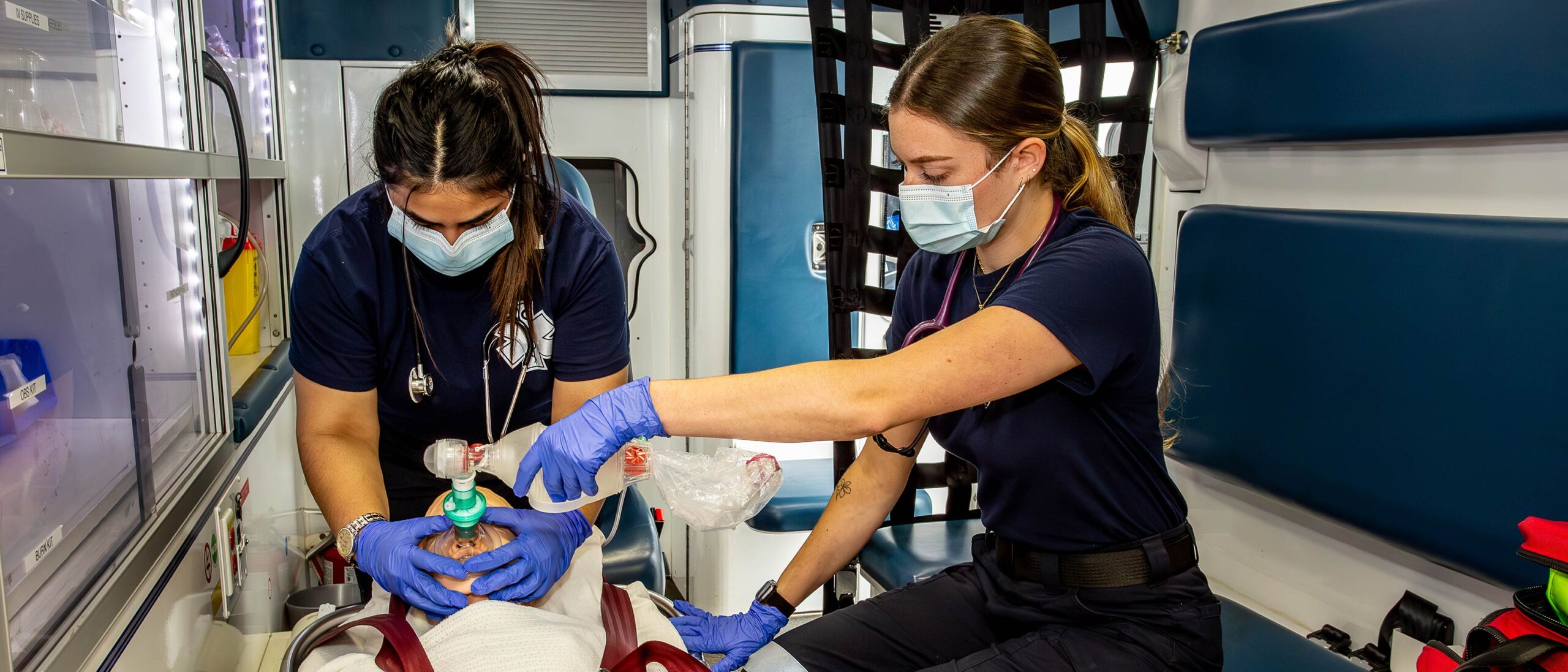 Two paramedic students are working with a mock patient inside an ambulance. The students are wearing professional attire and wearing masks.