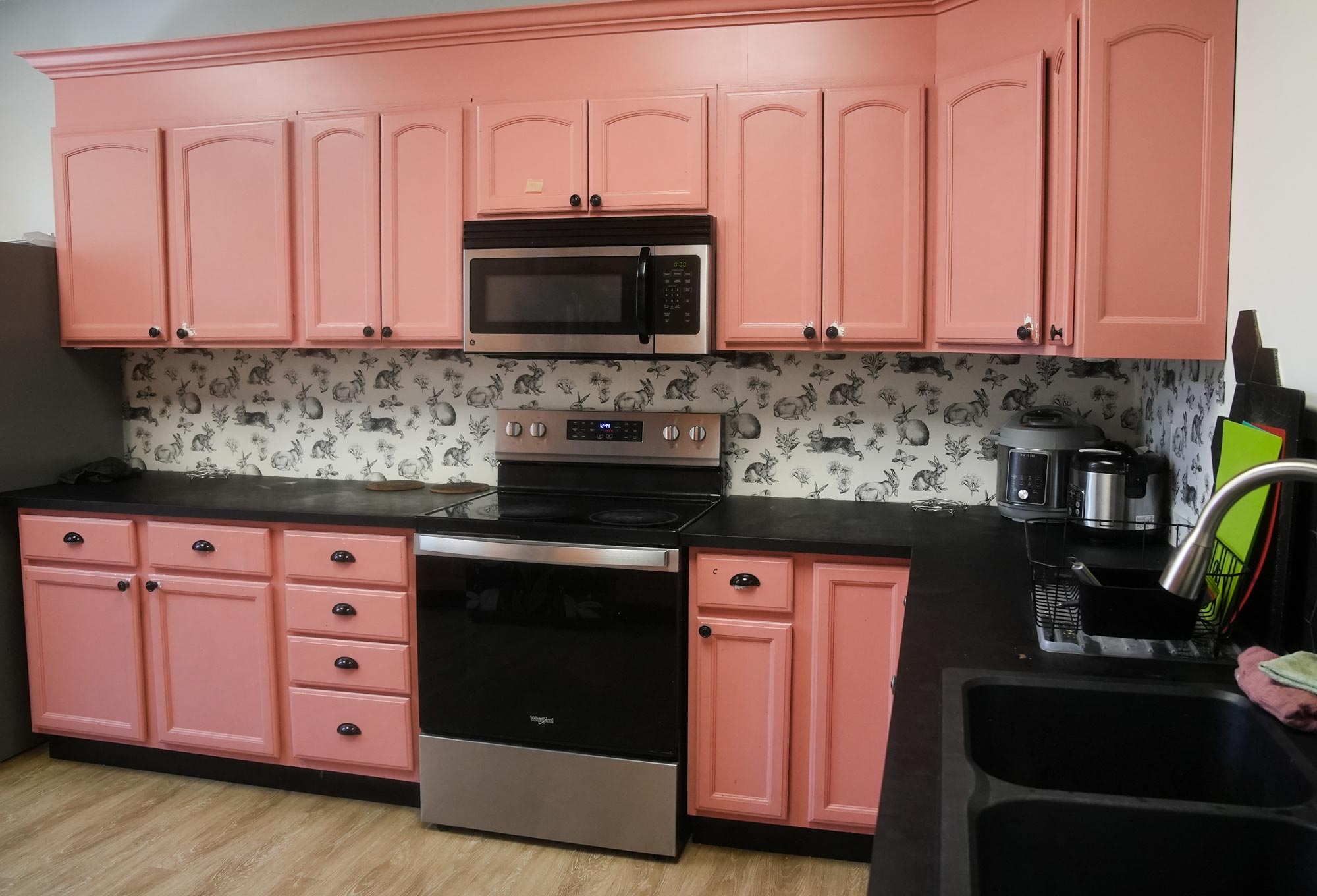 A kitchen with pink cabinets, microwave over a stove, various kitchen appliances and a sink area.