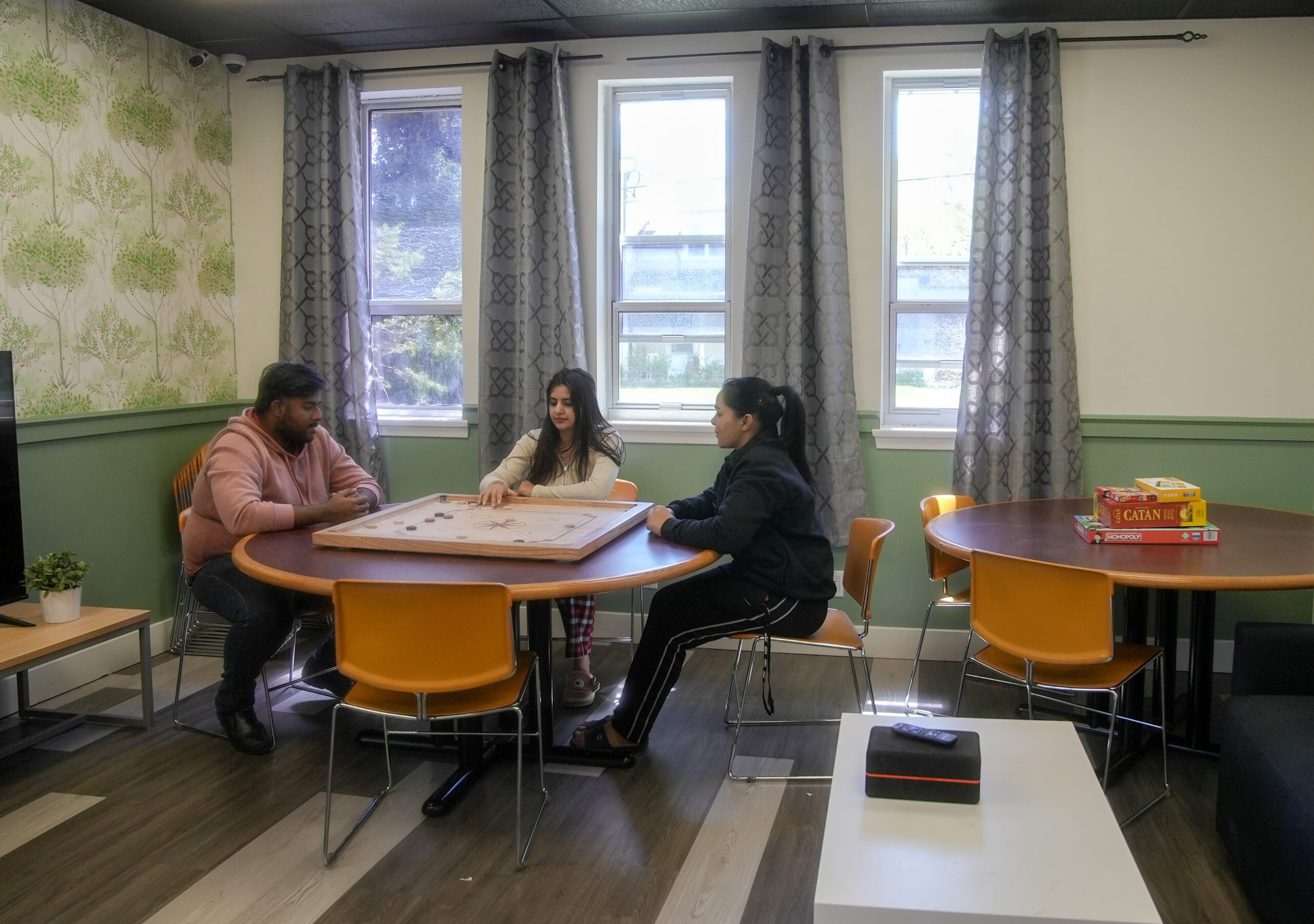 Students are sitting at a table playing a board game. The students are next to a set of windows with curtains. There is a table with a set of chairs beside them on the right with a set of boardgames on top.