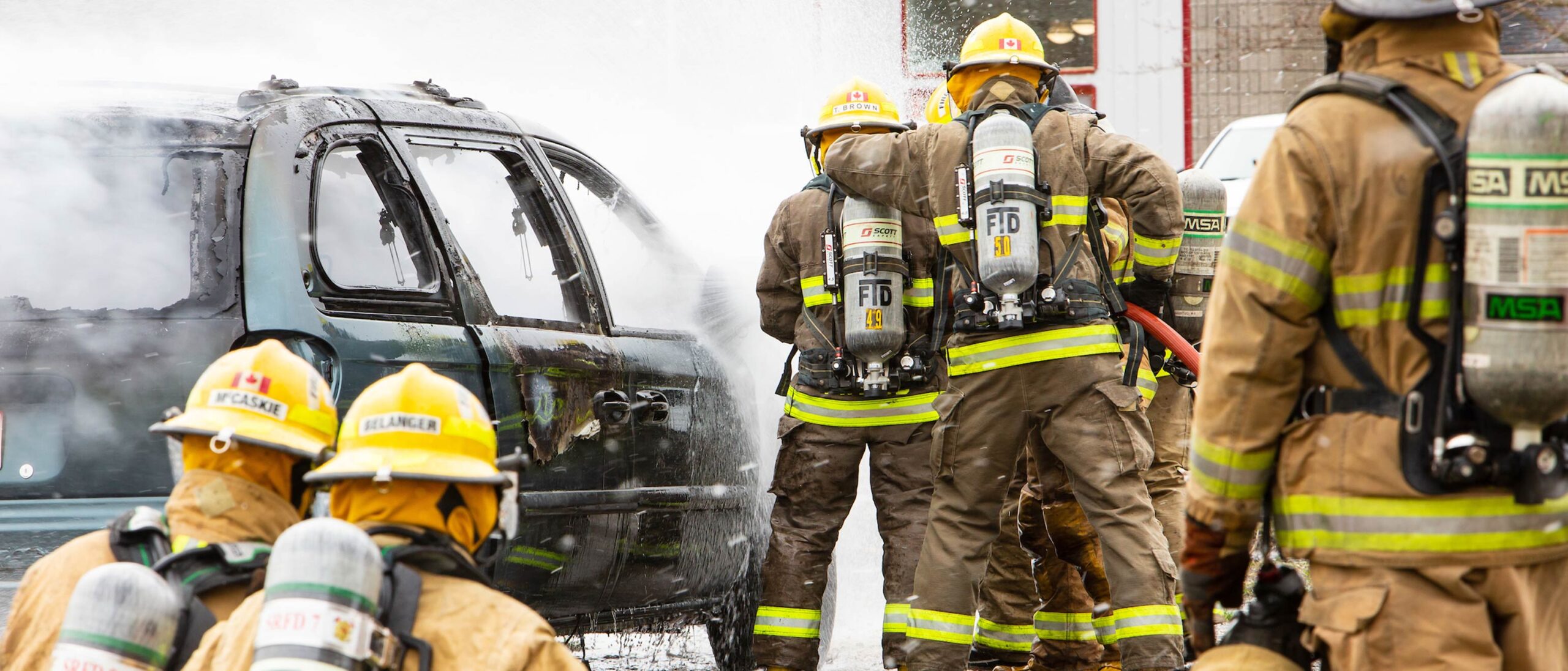 A group of students in the pre-service firefighter program are wearing professional gear and using appropriate attire to get a fire out from a car.