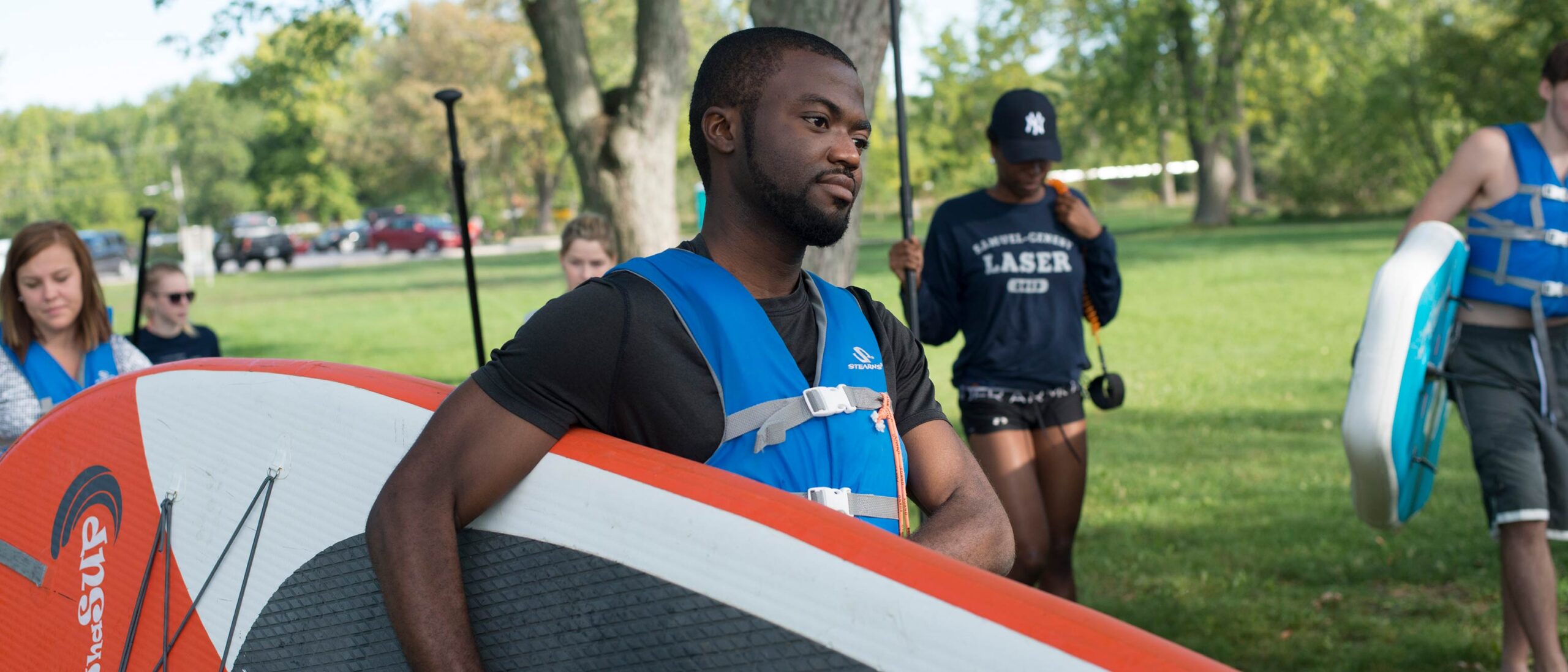 A student in a life jacket carries a paddle board under their arm, walking outside through a grassy field. Other students walk alongside and behind carrying paddles and other paddle boards.