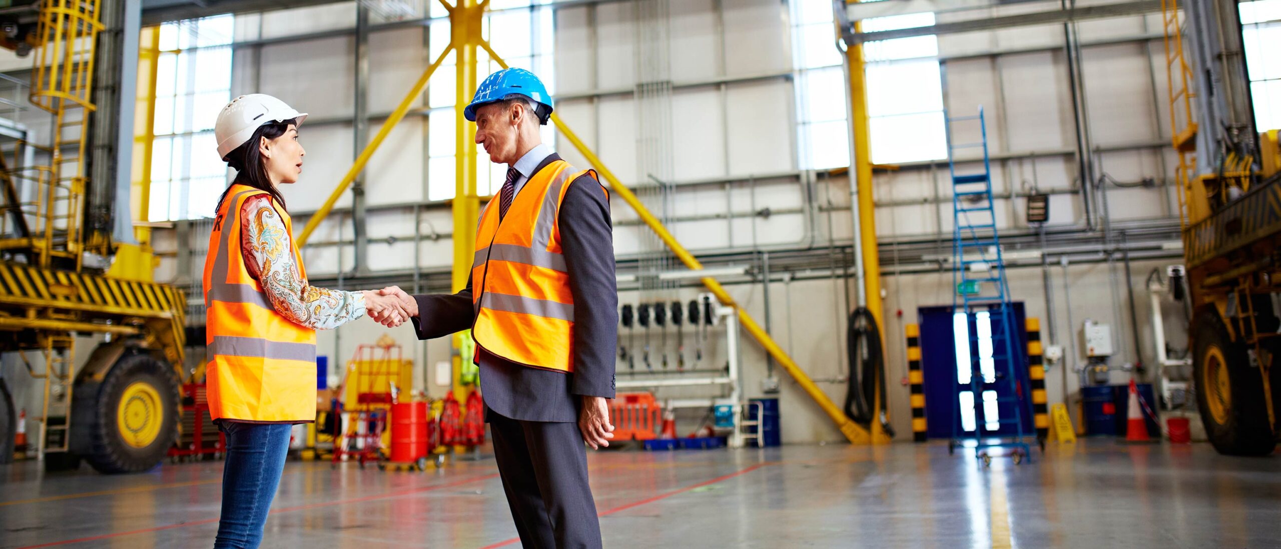 Two people wearing hard hats and high visibility vests shake hands in the middle of an industrial factory.