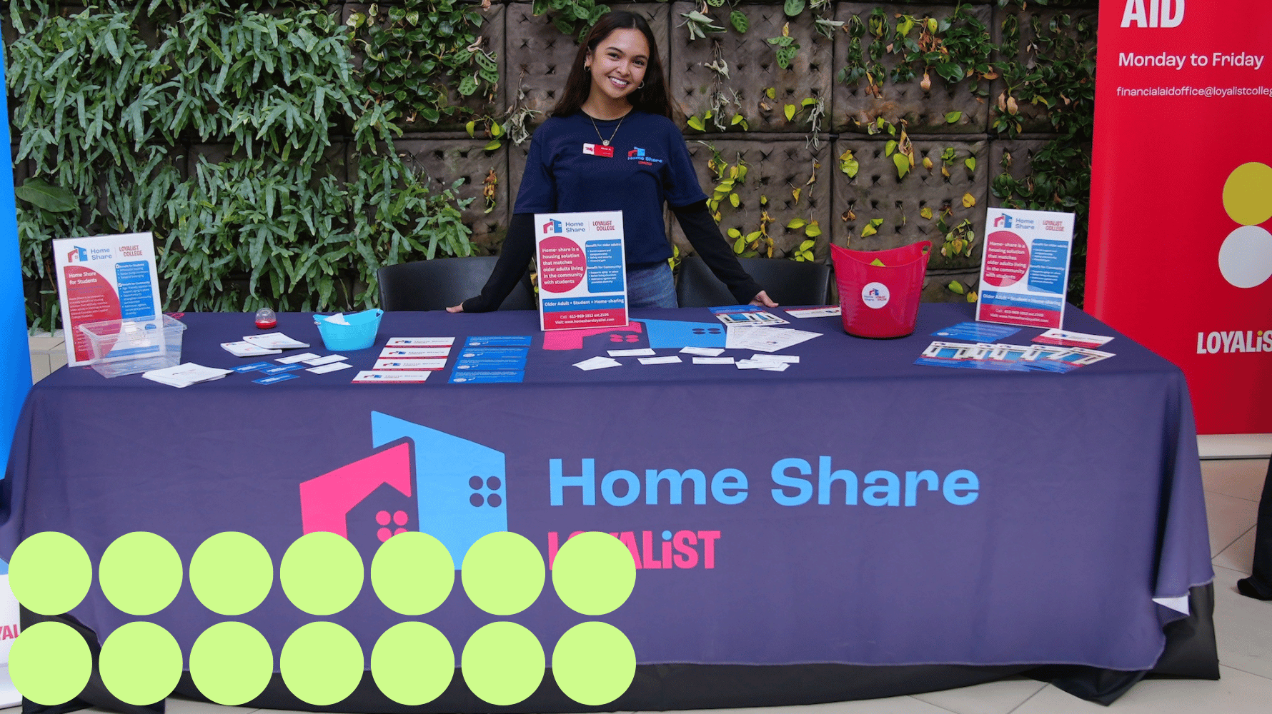 A student stands behind a table filled with informational brochures and cards. The tablecloth says "Home Share, Loyalist." Green graphic circles are in the lower lefthand corner of the image.