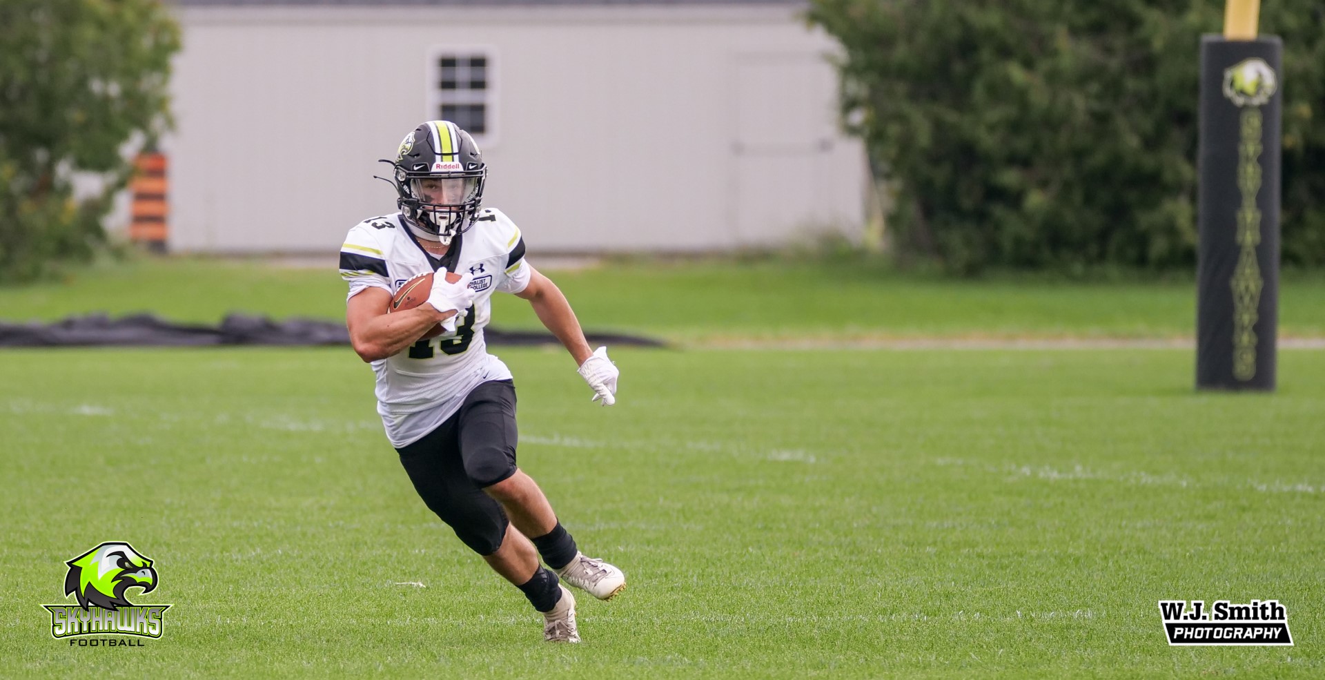 A Skyhawks football player runs over the field, holding a football in one hand.