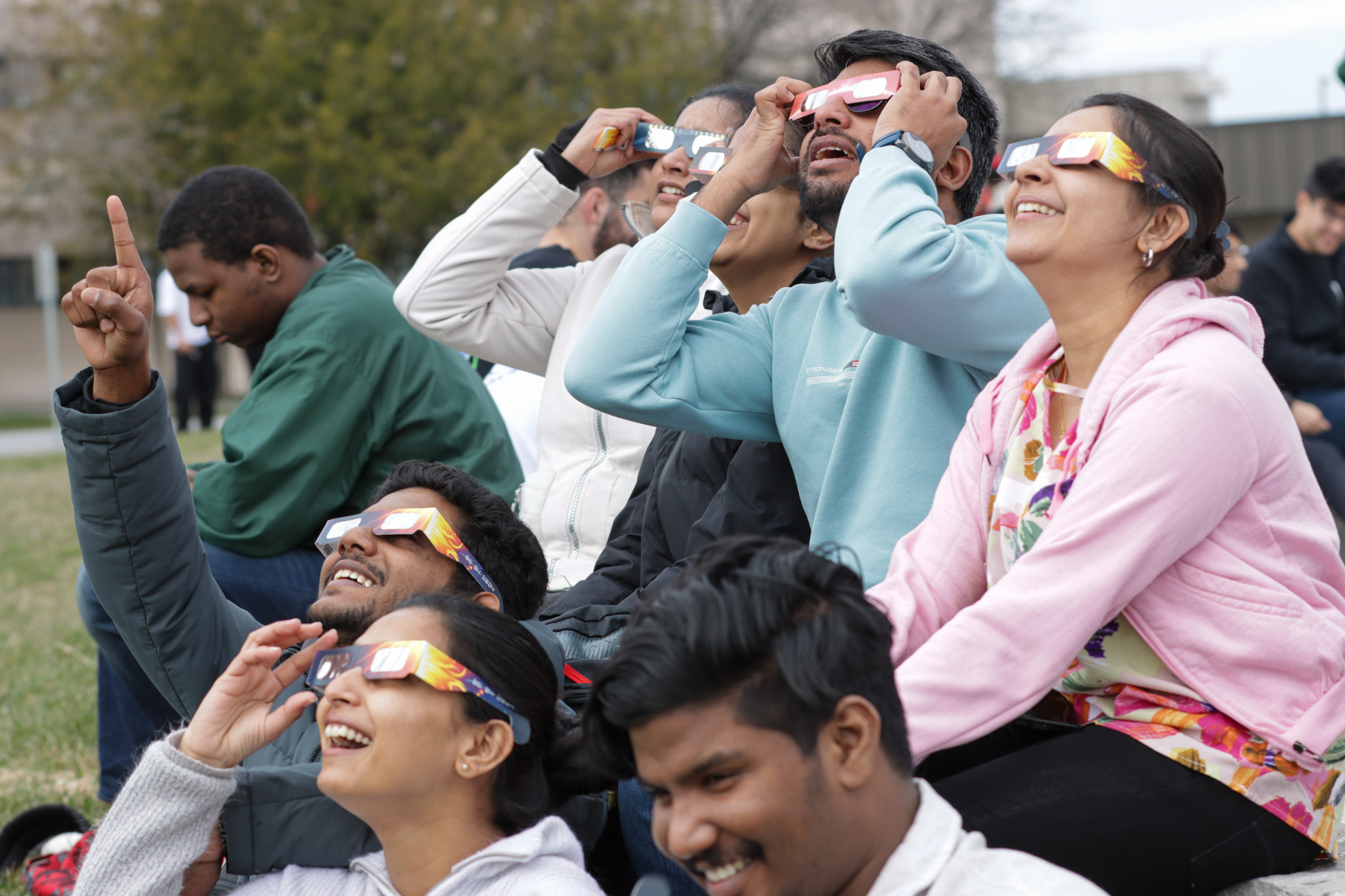 A group of students is gathered outdoors, enthusiastically watching the sky during a solar event. They are all wearing special solar viewing glasses, their faces lit up with excitement and curiosity. Some are pointing upwards, while others smile as they gaze at the phenomenon above. The diverse group sits closely together, sharing this awe-inspiring experience in a spirit of community and learning.