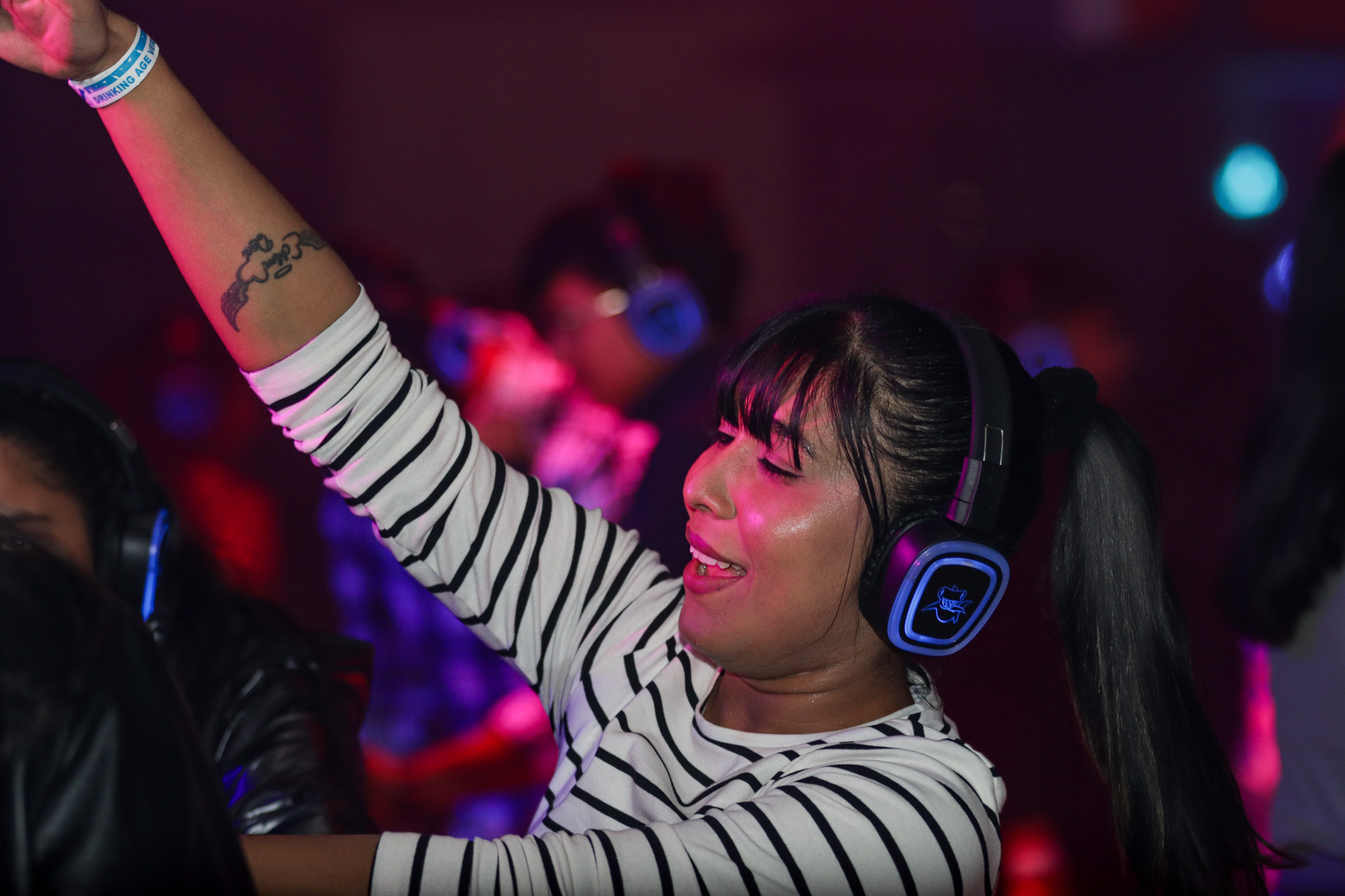 A person enjoying a silent disco, wearing headphones and dancing with enthusiasm. The vibrant lighting and striped shirt add to the lively atmosphere, as they raise an arm, fully immersed in the music.