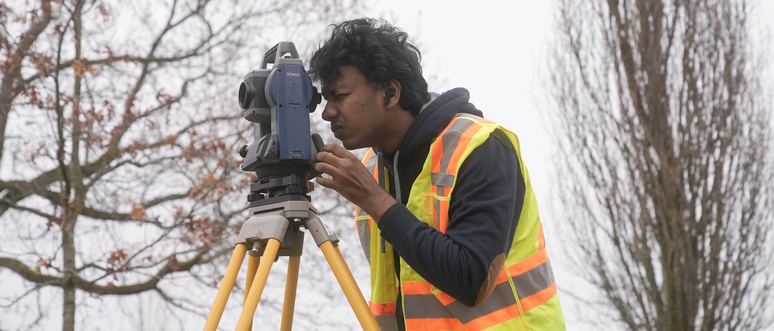 A student in a high visibility vest takes a measurement for a land survey, looking through the viewer in front of two trees.