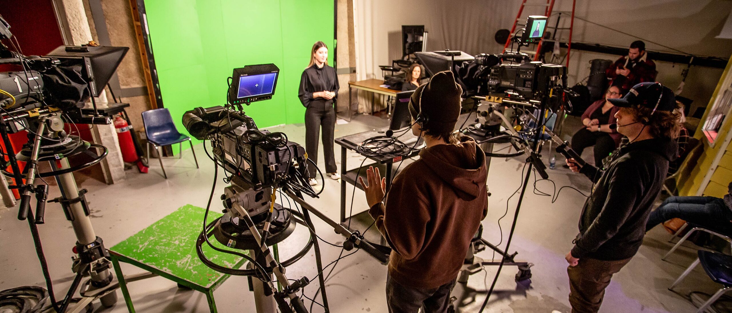 A student stands before a green screen in a television lab, where other students operate the cameras and monitor the feed.