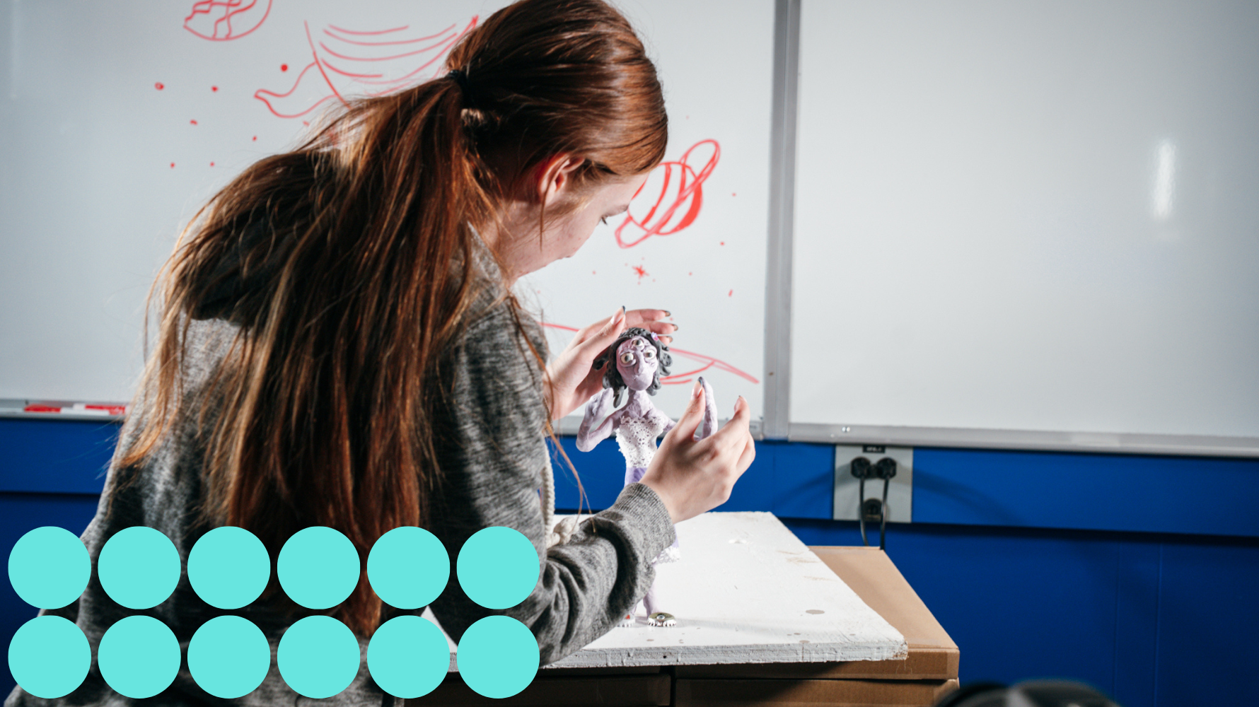 A student works on a clay animation figure in a studio classroom. The student carefully adjusts the alien character model on a white tabletop, with sketches of planets and space-themed drawings visible on a whiteboard behind them.