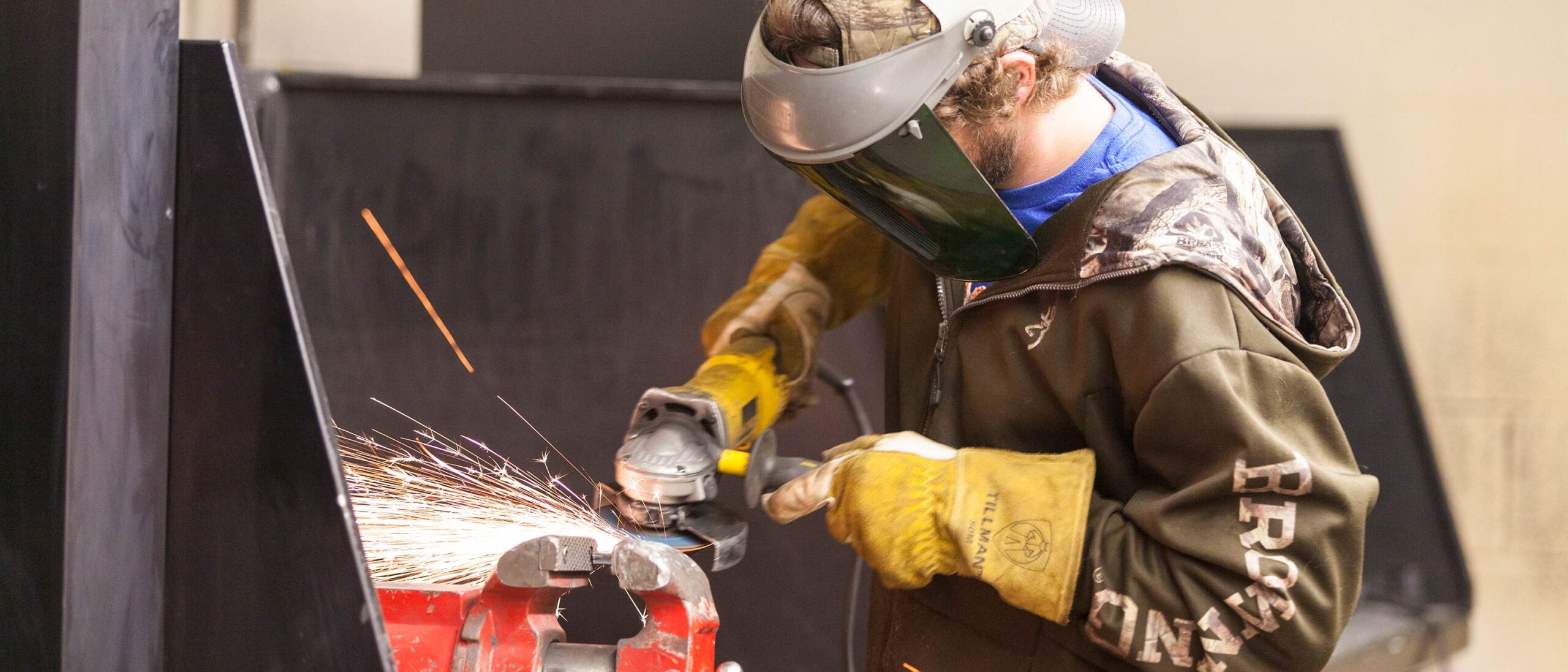 A student wearing a protective mask and gloves grinds a metal piece held in a vice. Blue graphic circles are in the lower lefthand corner of the image.