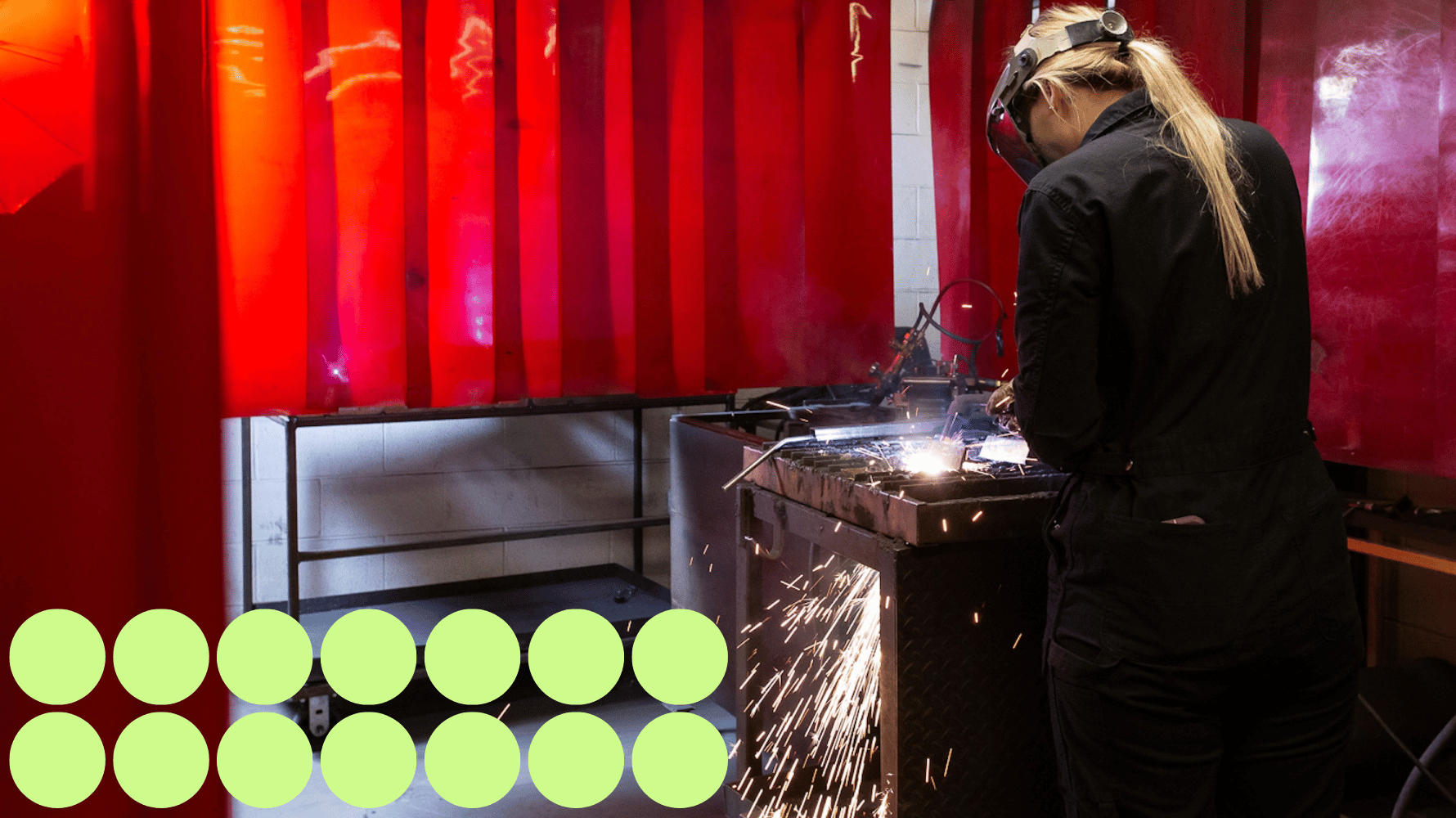 A student wearing a welding mask and gloves welds a workpiece on top of a metal bench in a welding booth with red shields. Green graphic circles are in the lower lefthand corner of the image.