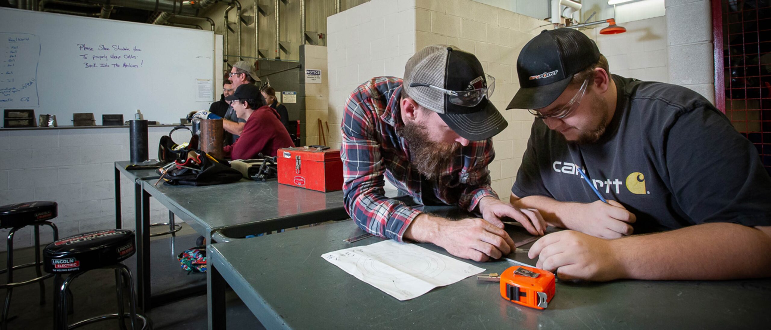 Two students work together to measure a metal part on a metal workbench, with a drawing and tape measure on top. Another group of students and a professor work at a bench in the background.