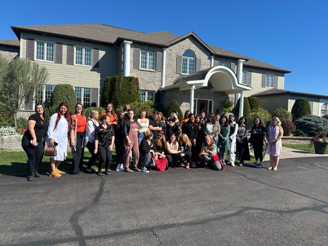 A group of second-year Esthetics students pictured with Catherine Wood and other staff in front of the Mahogany Salon and Spa in Carleton Place. The building in the background is a large home, with columns and green shrubs in front.