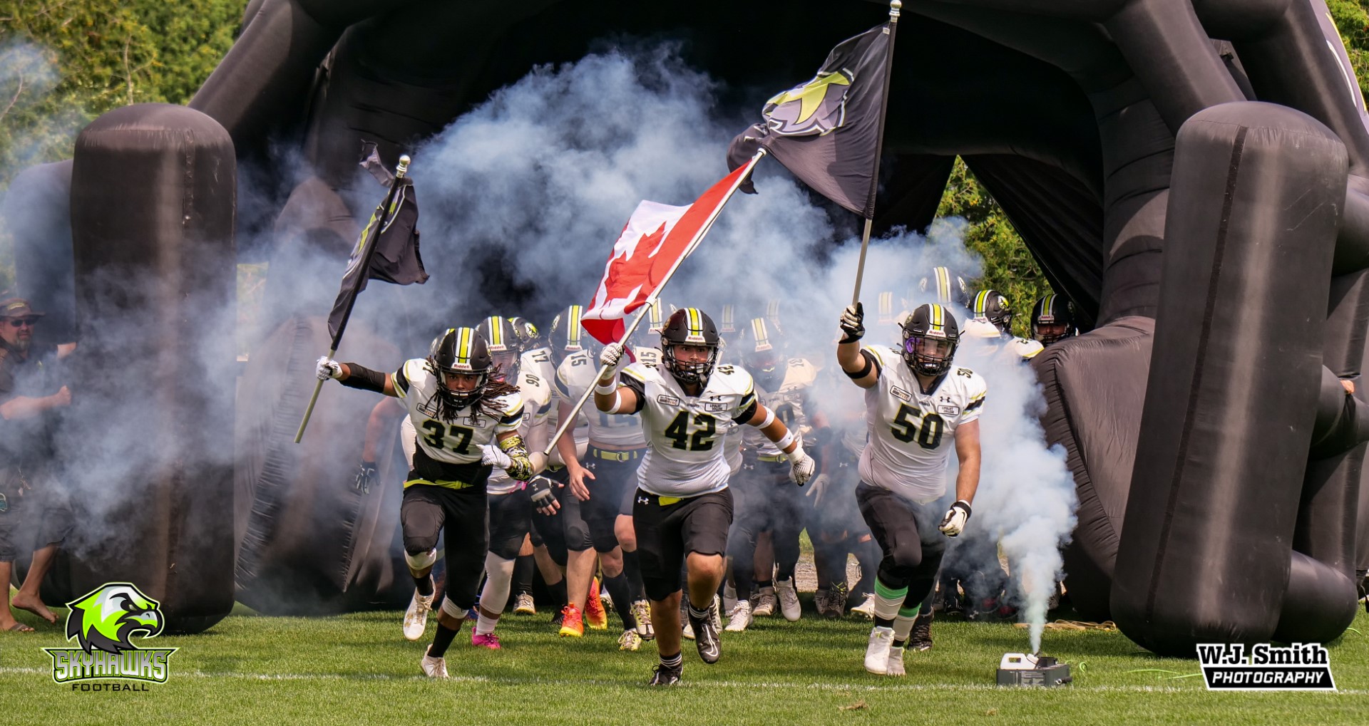 The Skyhawks football team runs out onto the field in their team equipment and jerseys. The front three are carrying two team flags and a Canadian flag. A smoke machine is placed where the students run out.