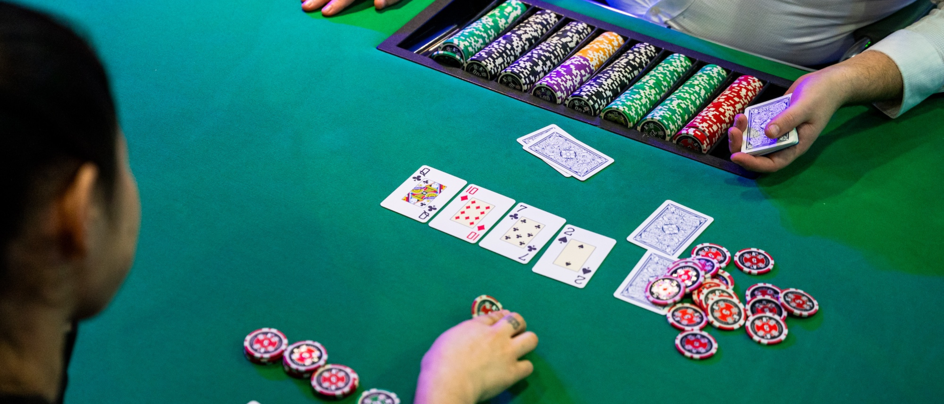 A casino table with a green felt surface where a poker game is in progress. A dealer hands out cards, and chips are stacked in the centre of the table. The focus is on a pair of Queen of Clubs and Ten of Diamonds on the table, and a few poker chips in a player’s hand.